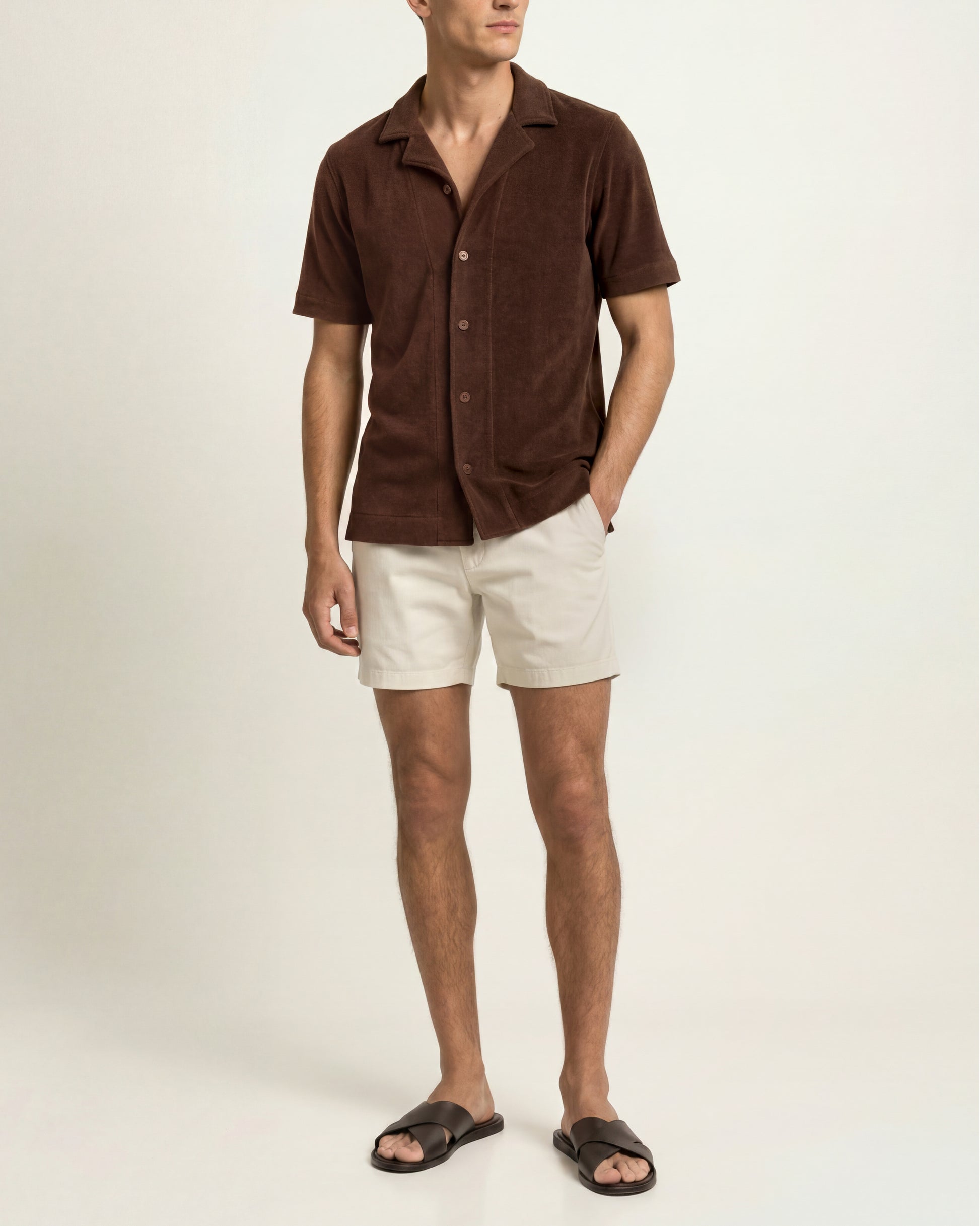 A man modeling a short-sleeve brown button-up shirt with cream shorts and black slide sandals, standing against a plain off-white studio background.