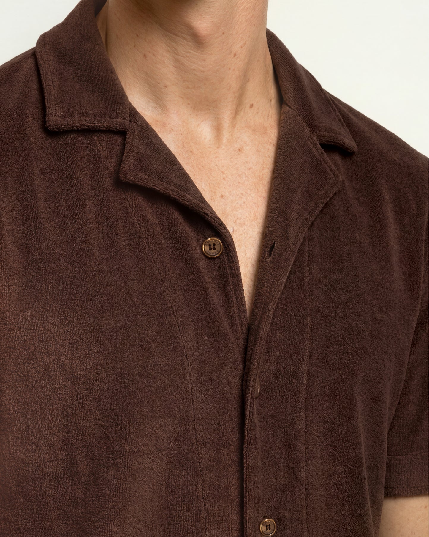 Close-up of a person wearing a short-sleeve button-front collared shirt in dark brown textured fabric, with the collar and upper chest visible against a plain light background.
