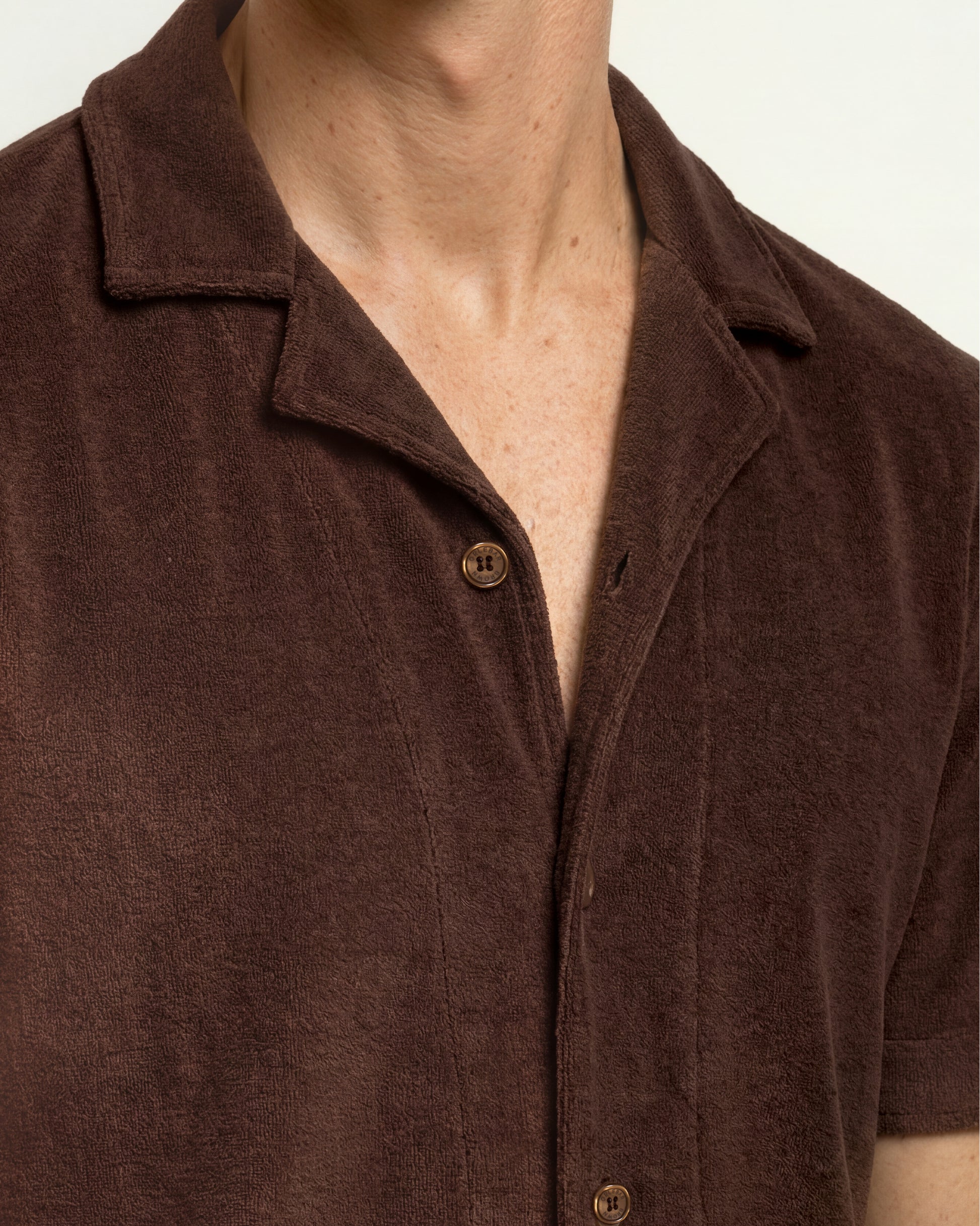 Close-up of a person wearing a short-sleeve button-front collared shirt in dark brown textured fabric, with the collar and upper chest visible against a plain light background.