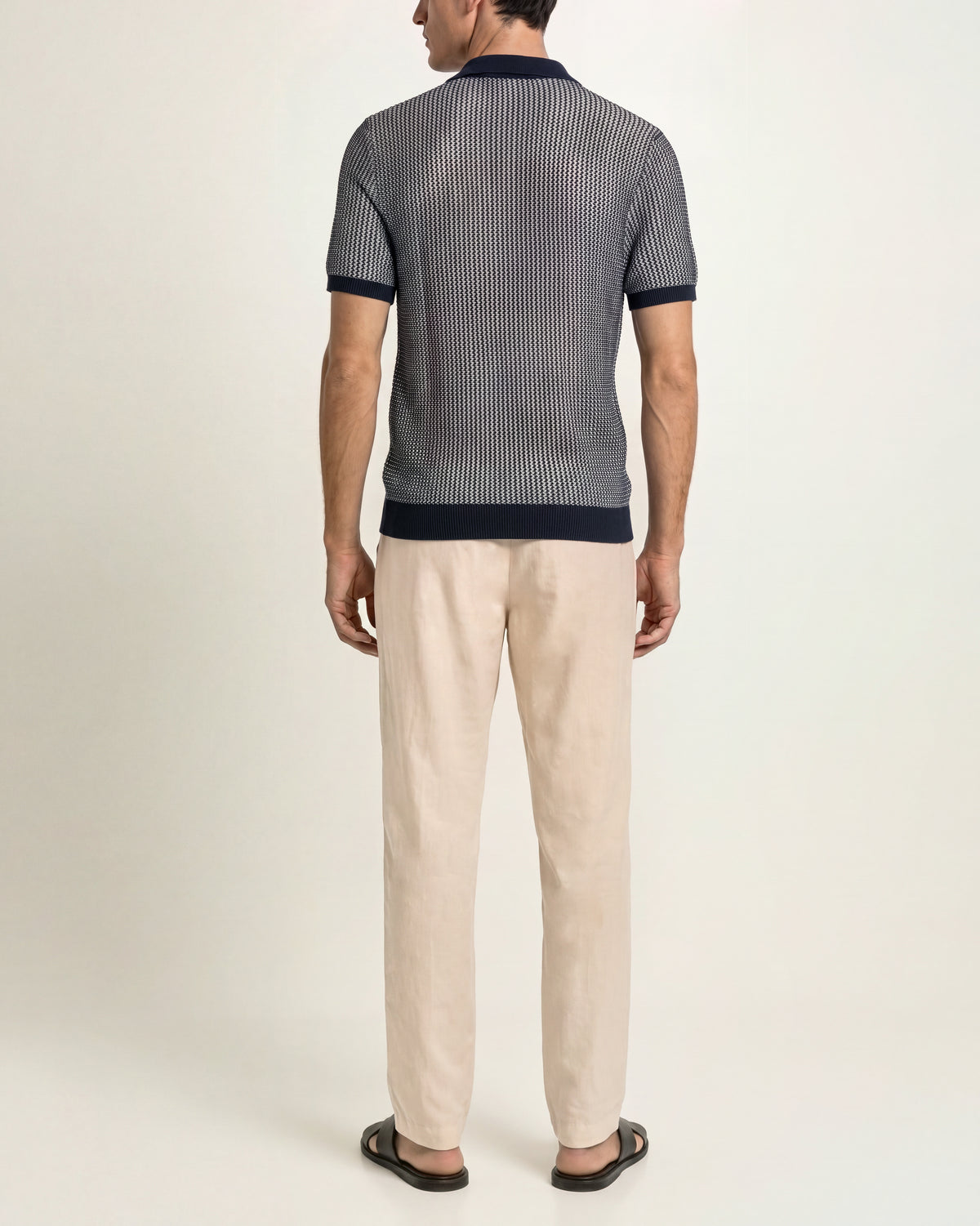 A man viewed from the back wearing a short-sleeve knit polo shirt with vertical navy and gray stripes, paired with beige trousers and dark sandals, against a plain light background.