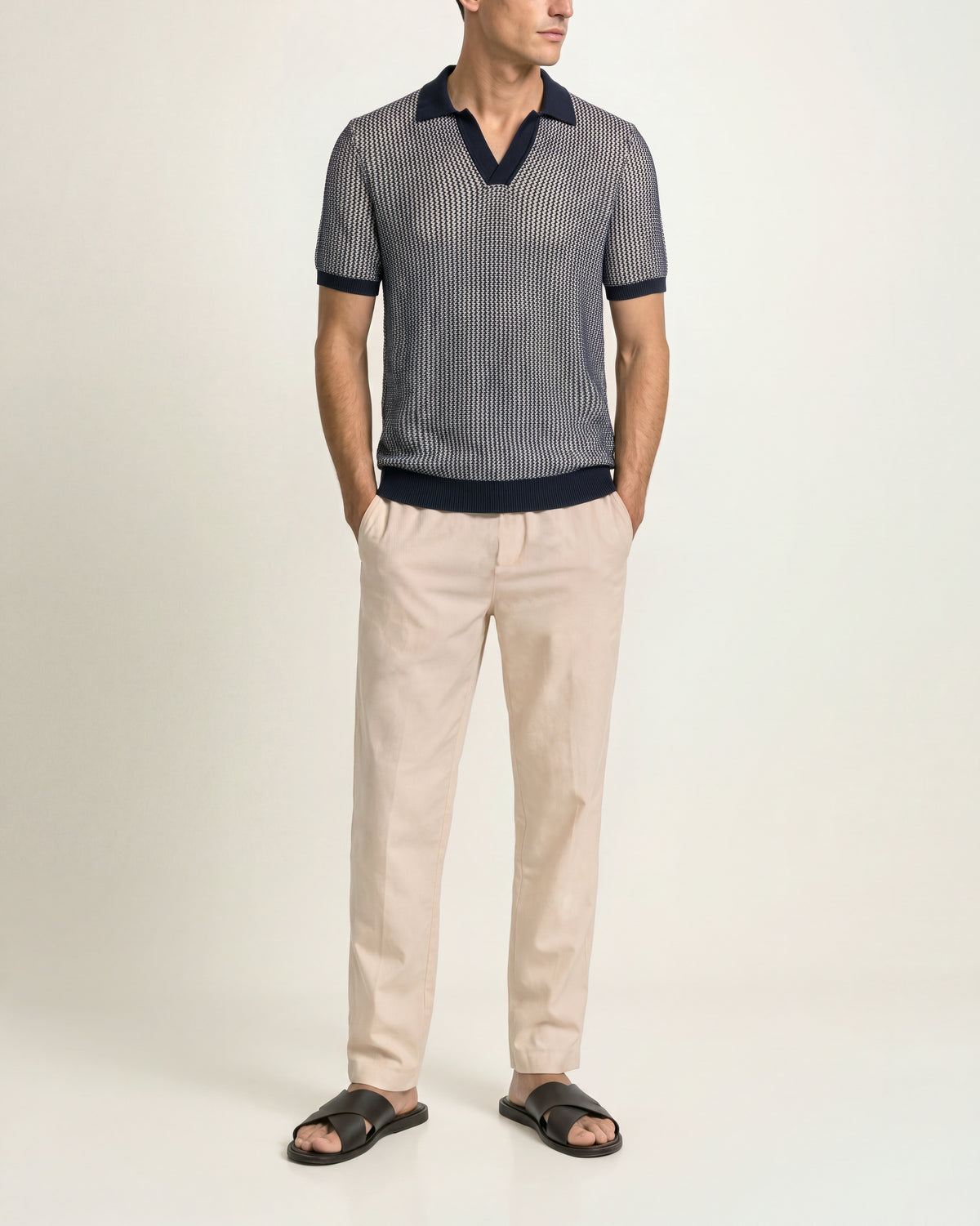 A man modeling a short-sleeve knit polo shirt with vertical gray and navy stripes, paired with beige trousers and black slide sandals, against a plain off-white studio background.