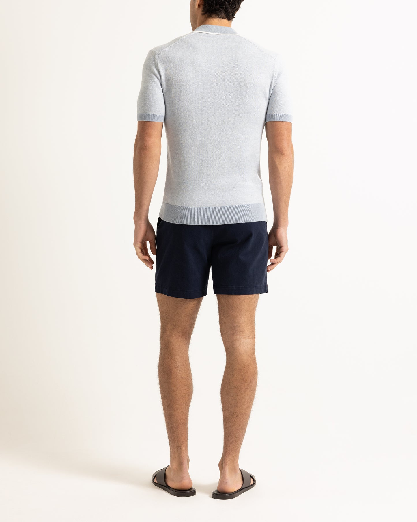 Man wearing light blue knitted polo shirt with navy shorts on a plain background