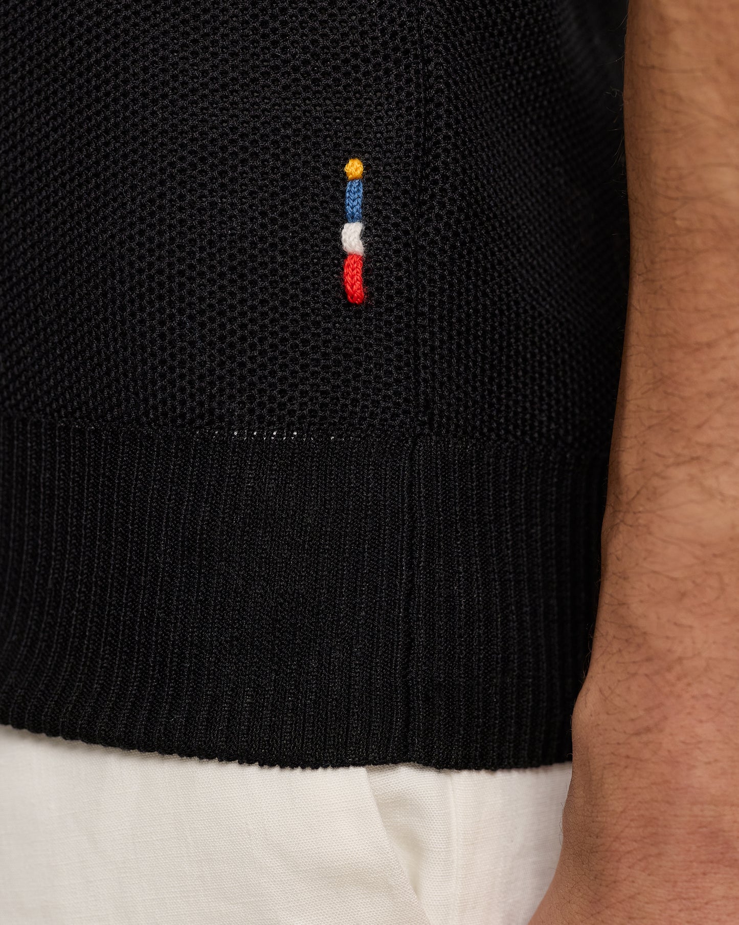 Black sweater with a colorful embroidered design on a person wearing white pants.