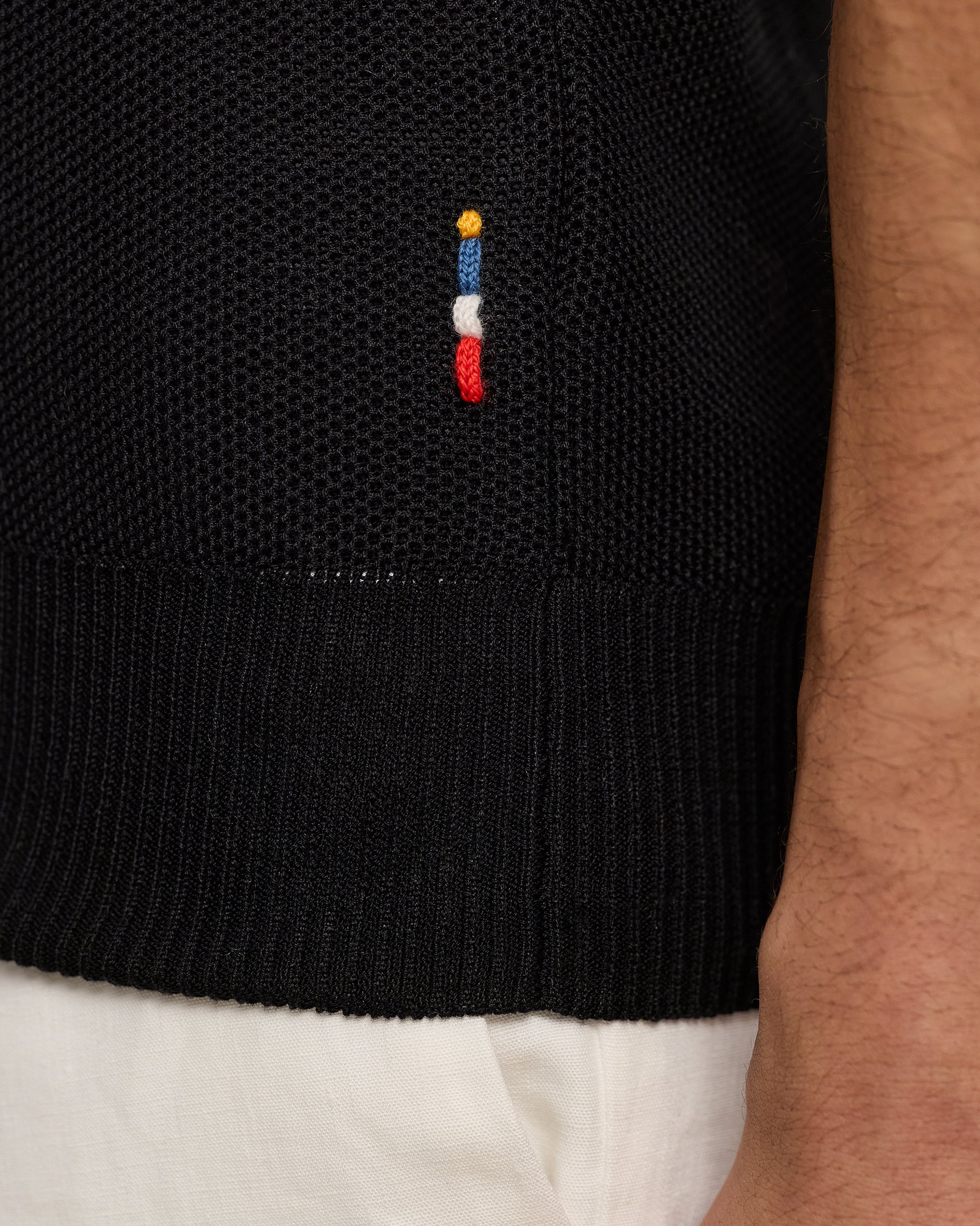 Black sweater with a colorful embroidered design on a person wearing white pants.
