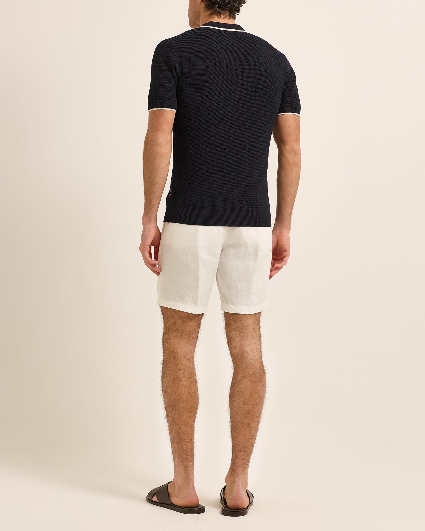 Man wearing navy polo shirt with white trim detailing and white shorts on a plain background