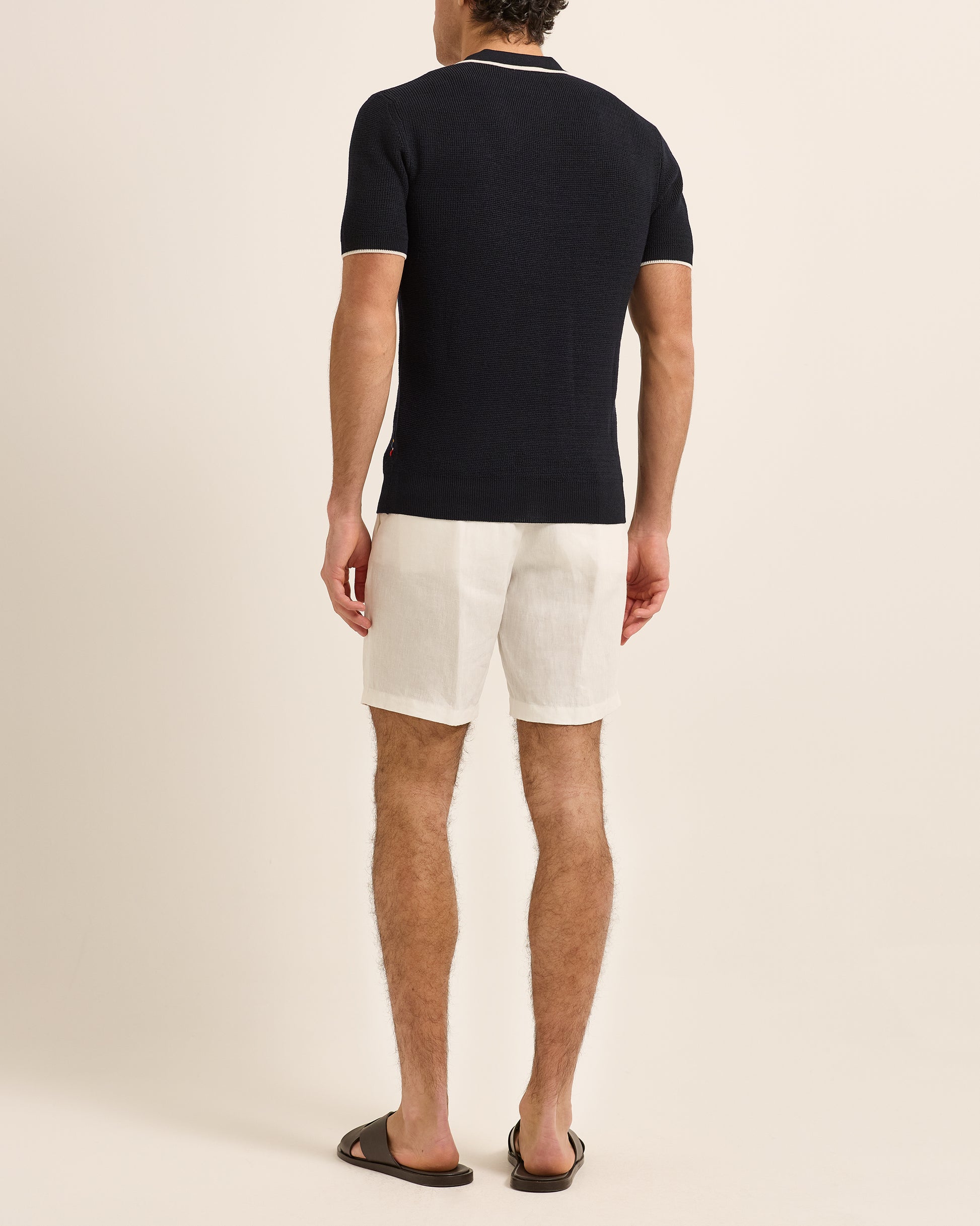 Man wearing navy polo shirt with white trim detailing and white shorts on a plain background