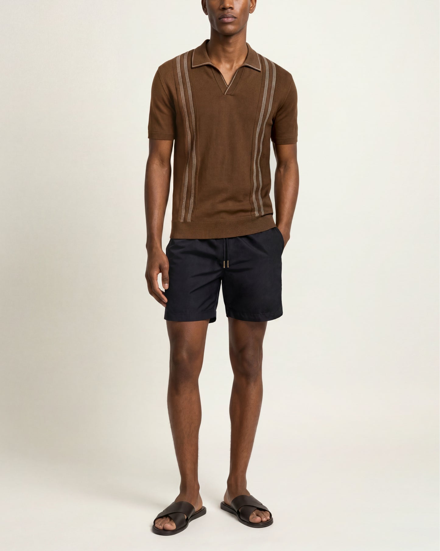 A man modeling a brown short-sleeve knit polo shirt with vertical beige stripe details on the chest, paired with dark navy shorts and dark brown slide sandals, against a plain light beige studio background.