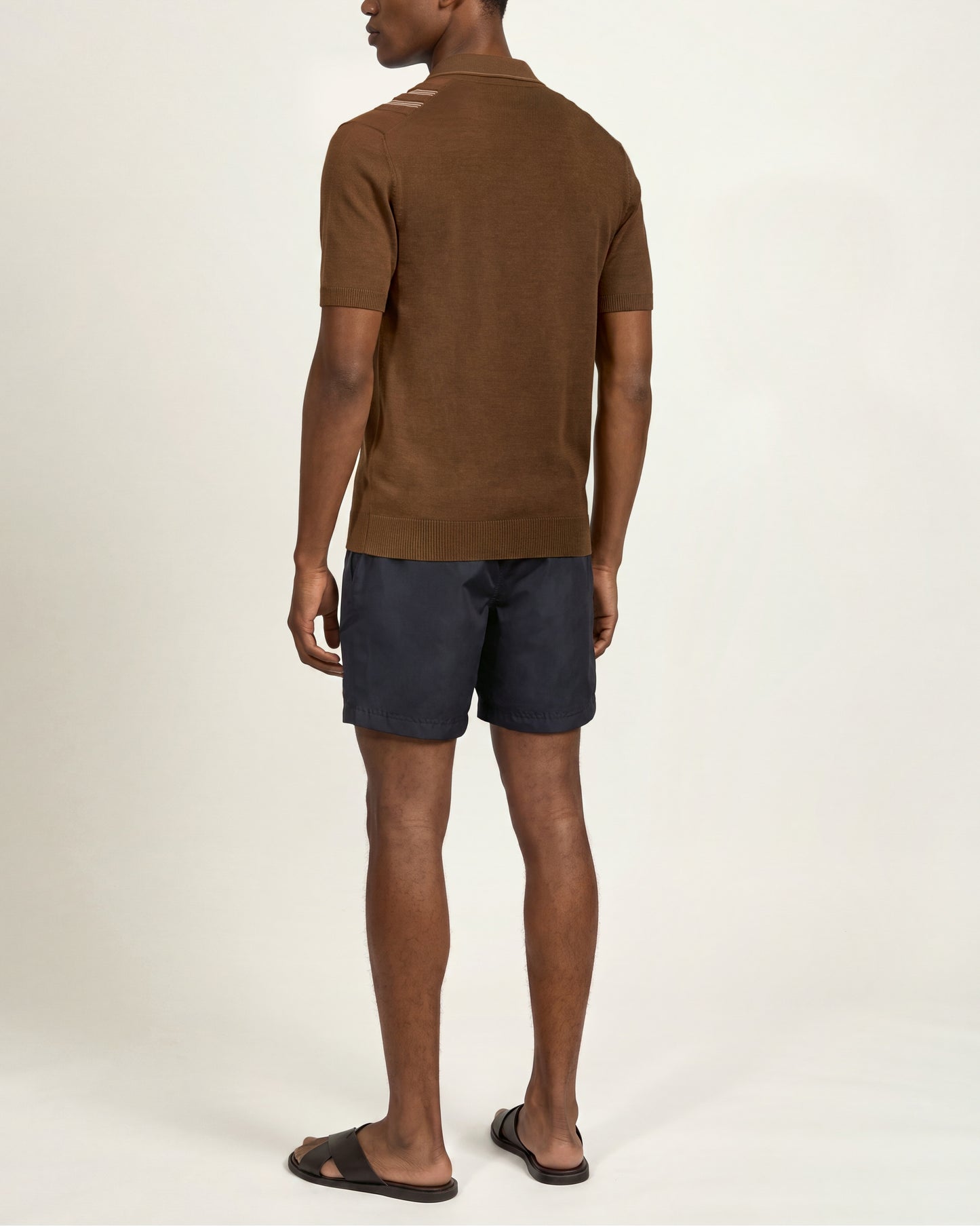 A man shown from the back wearing a brown short-sleeve knit top with details at the shoulders, navy shorts, and dark sandals, against a plain off-white studio background.