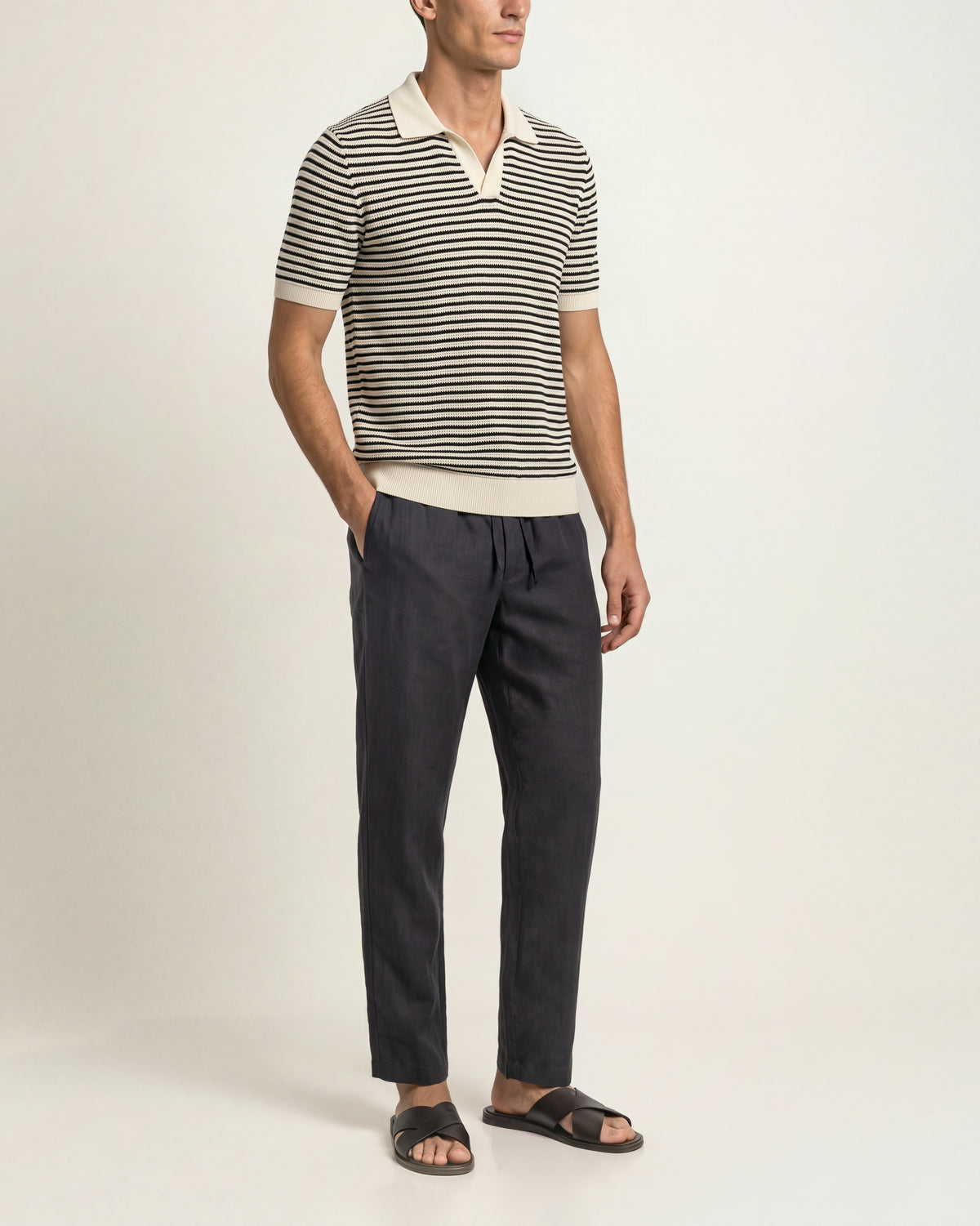 A man wearing a short-sleeve knit polo shirt with thin black and off-white horizontal stripes, paired with dark charcoal trousers and black slide sandals, standing against a plain light beige studio background.