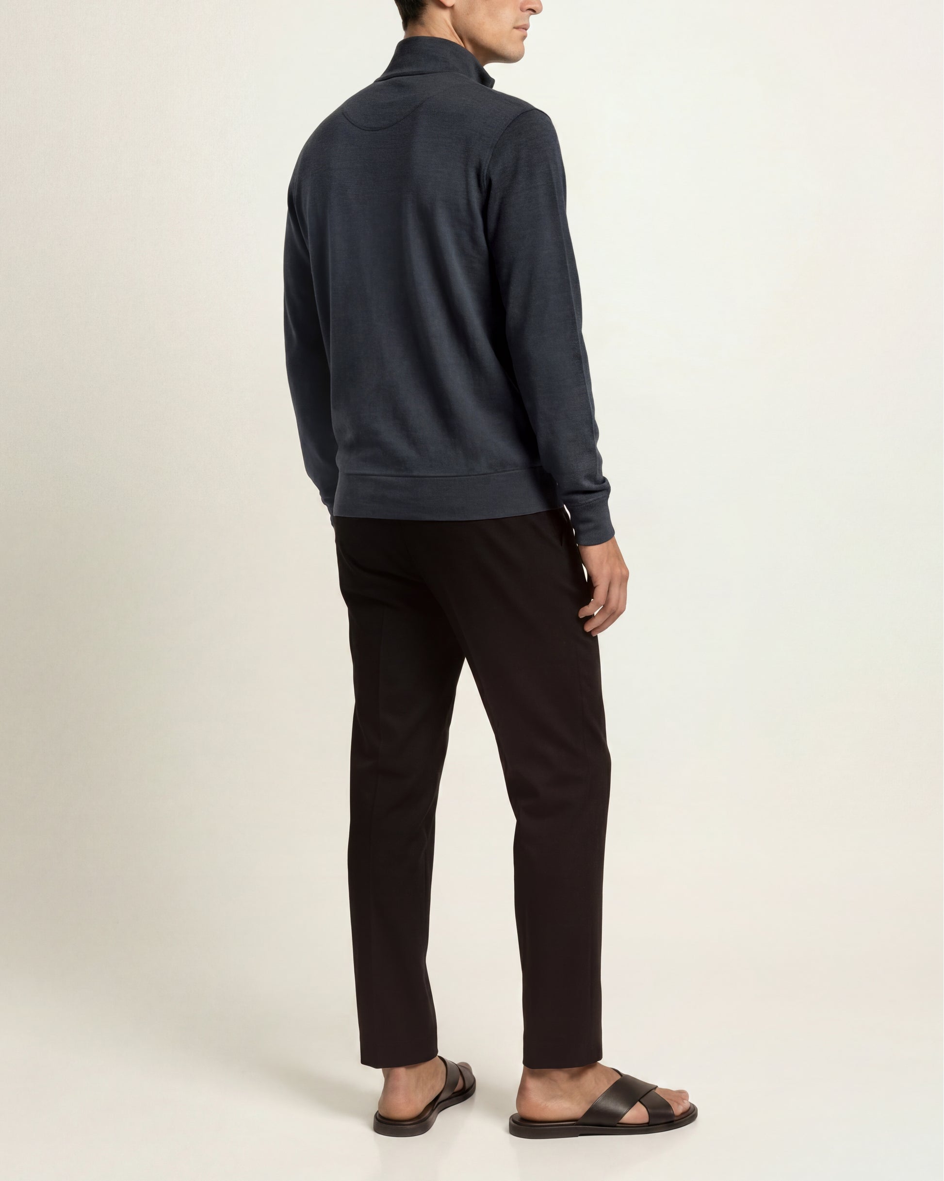 A man shown from the back wearing a dark navy quarter-zip pullover, dark brown trousers, and dark brown slide sandals, photographed against a plain off-white studio background.