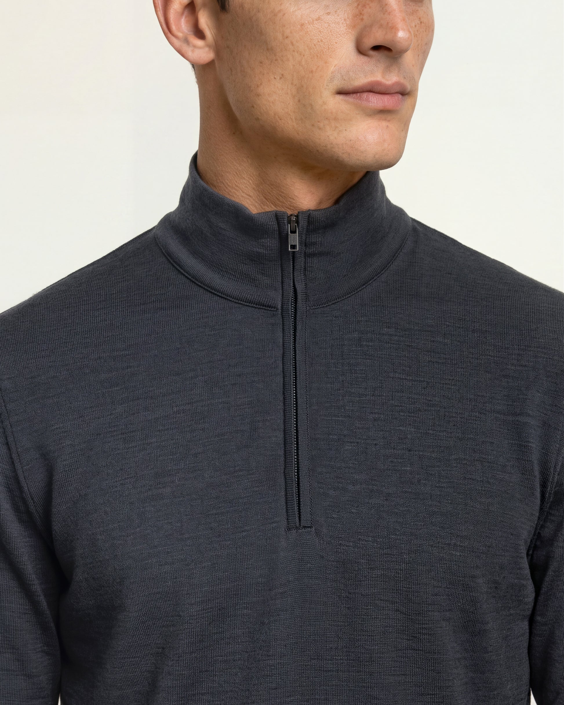 Close-up of a man wearing a dark charcoal gray quarter-zip mock neck pullover against a light off-white background.