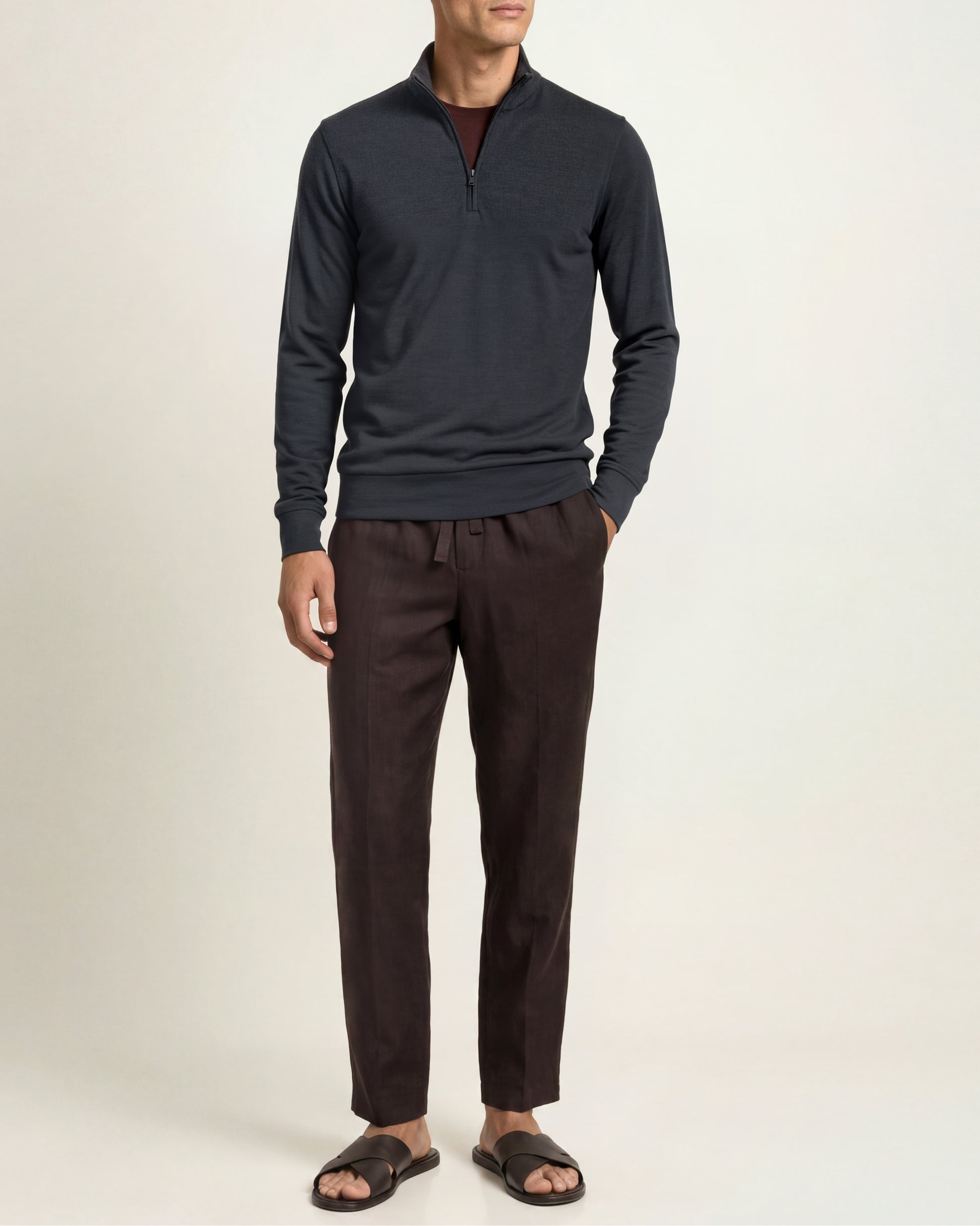A man modeling a dark charcoal gray quarter-zip long-sleeve shirt and dark brown drawstring trousers, paired with dark brown slide sandals, against a plain off-white studio background.