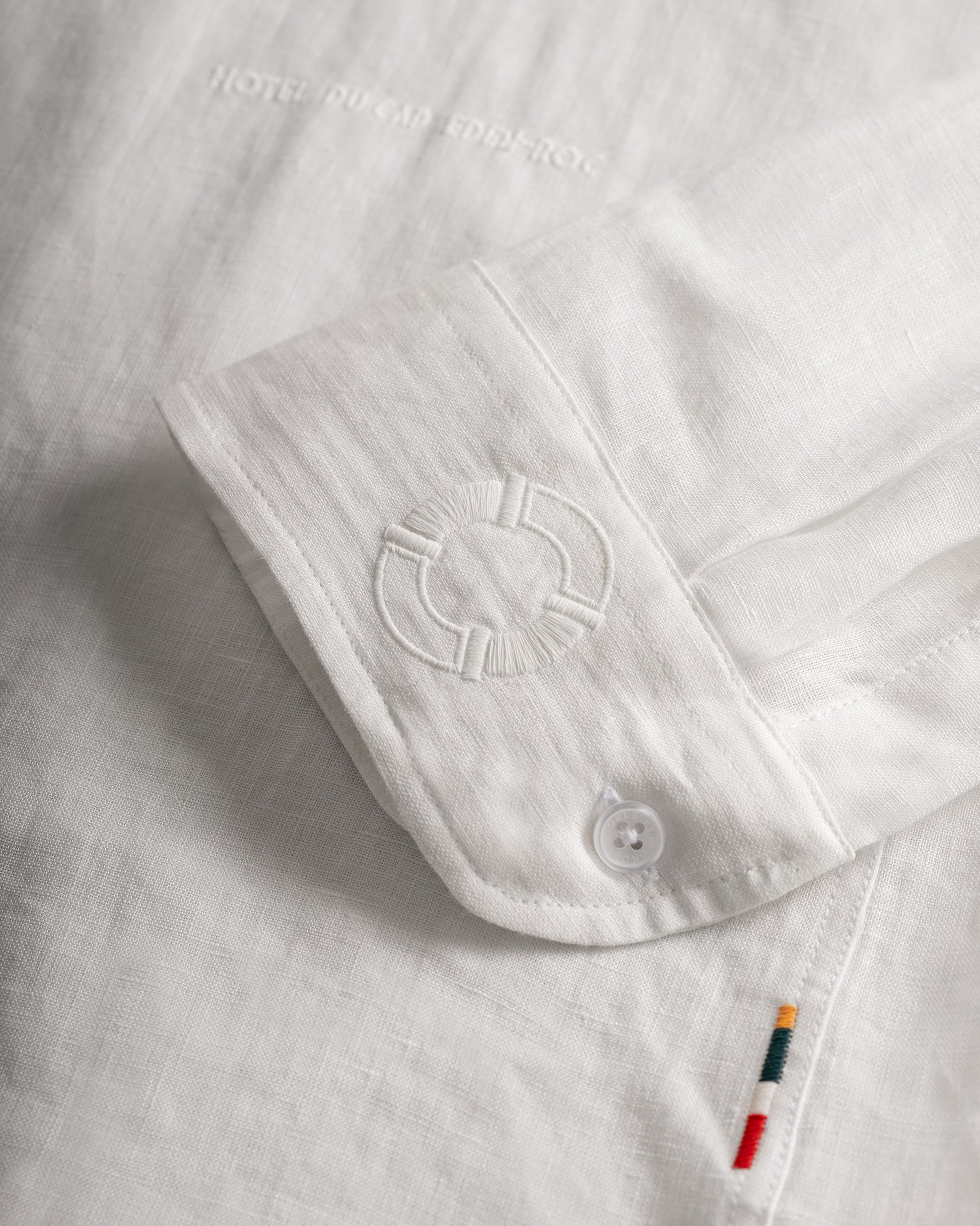Close-up of a white linen shirt sleeve with a buttoned cuff, tonal embroidered logo, and a small colorful striped accent tab near the hem.