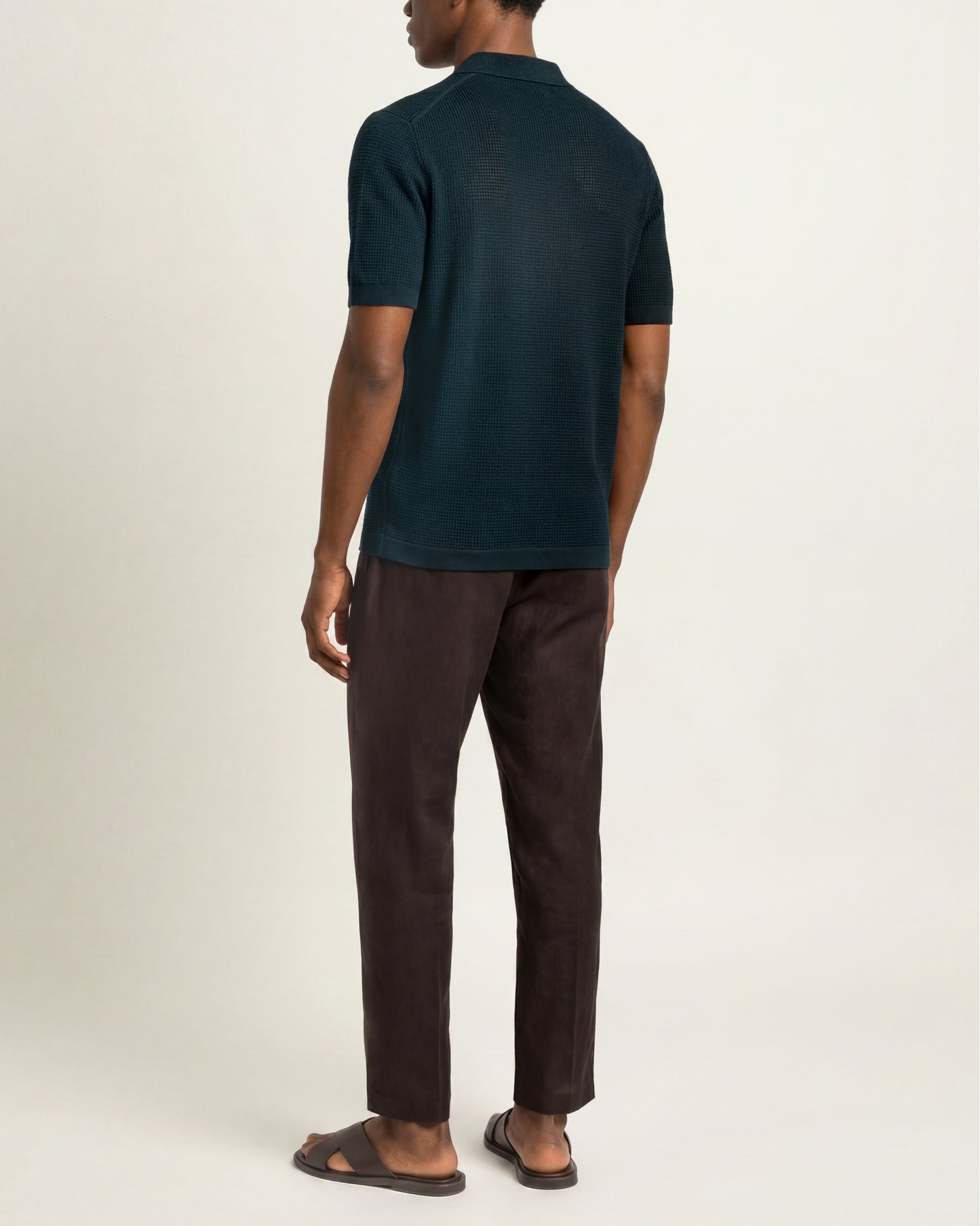 A man shown from the back wearing a dark teal short-sleeve polo shirt, dark brown trousers, and brown sandals against a plain off-white studio background.