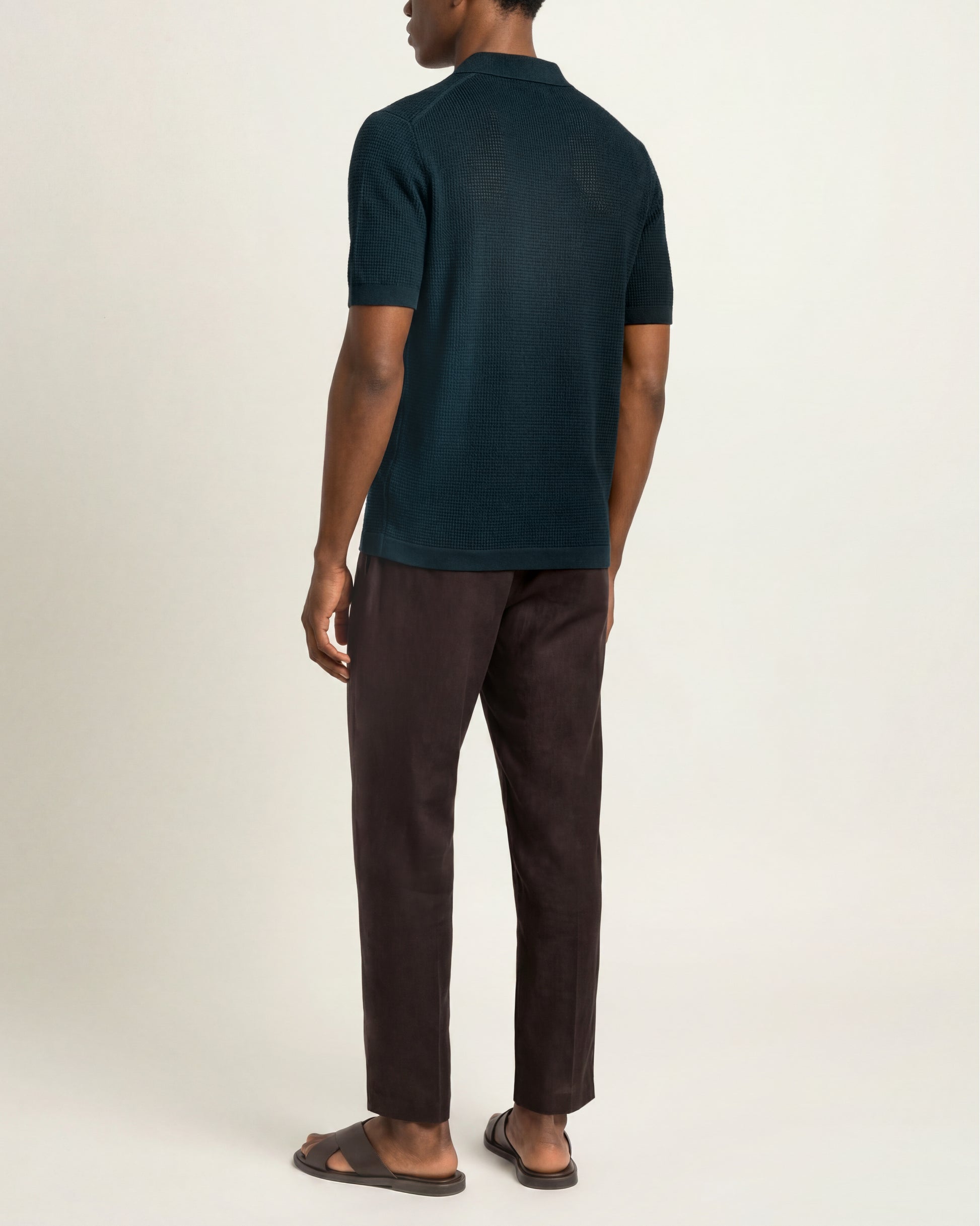 A man shown from the back wearing a dark teal short-sleeve polo shirt, dark brown trousers, and brown sandals against a plain off-white studio background.