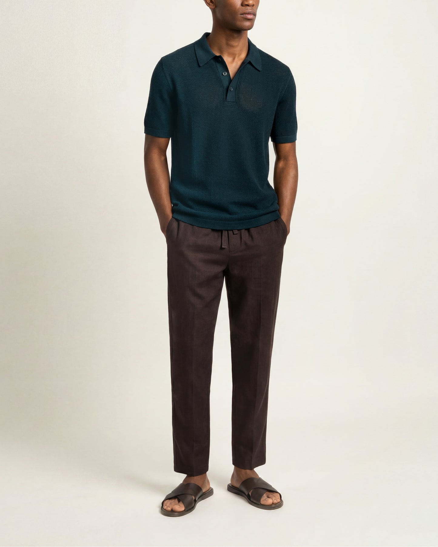 A man wearing a dark teal short-sleeve polo shirt and dark brown drawstring trousers, paired with dark brown slide sandals, standing against a plain off-white studio background.