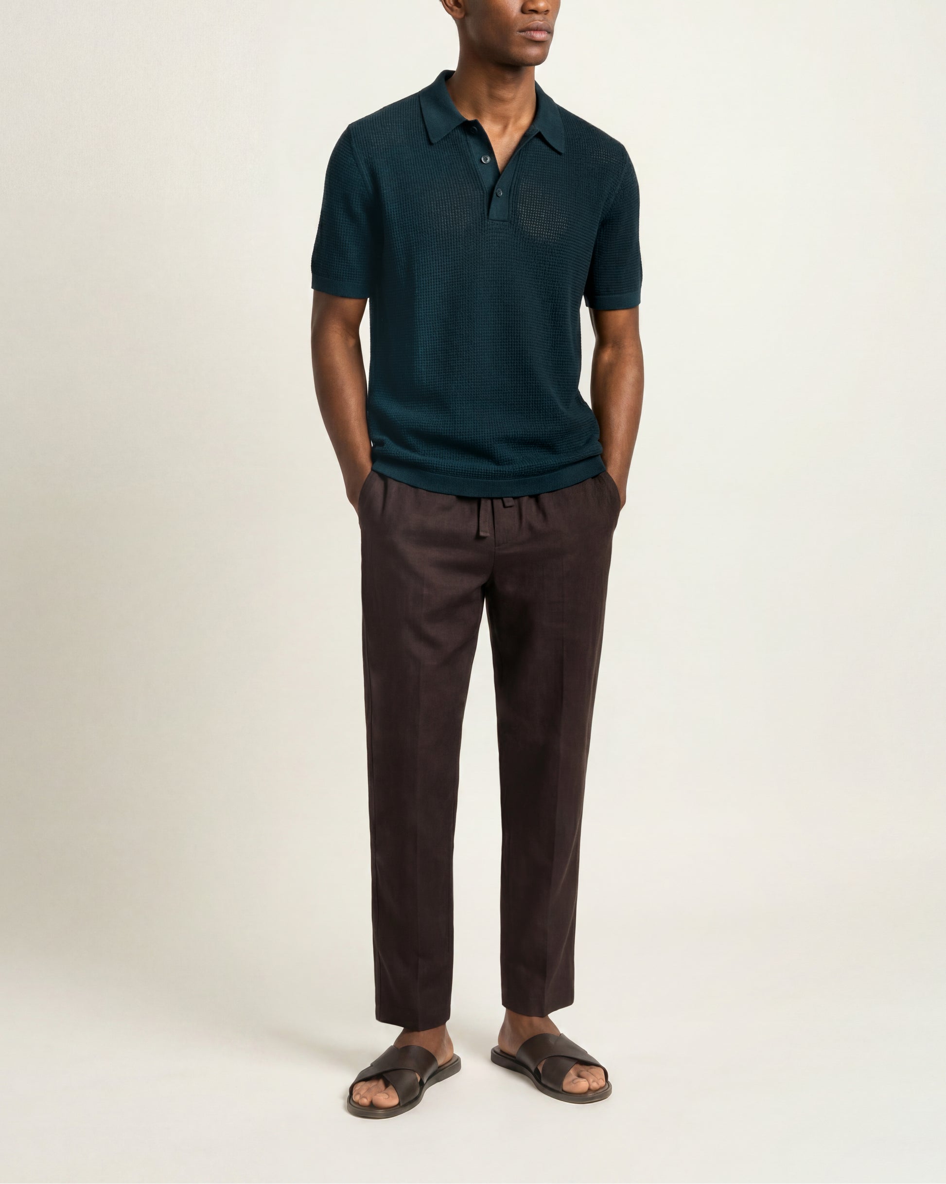A man wearing a dark teal short-sleeve polo shirt and dark brown drawstring trousers, paired with dark brown slide sandals, standing against a plain off-white studio background.