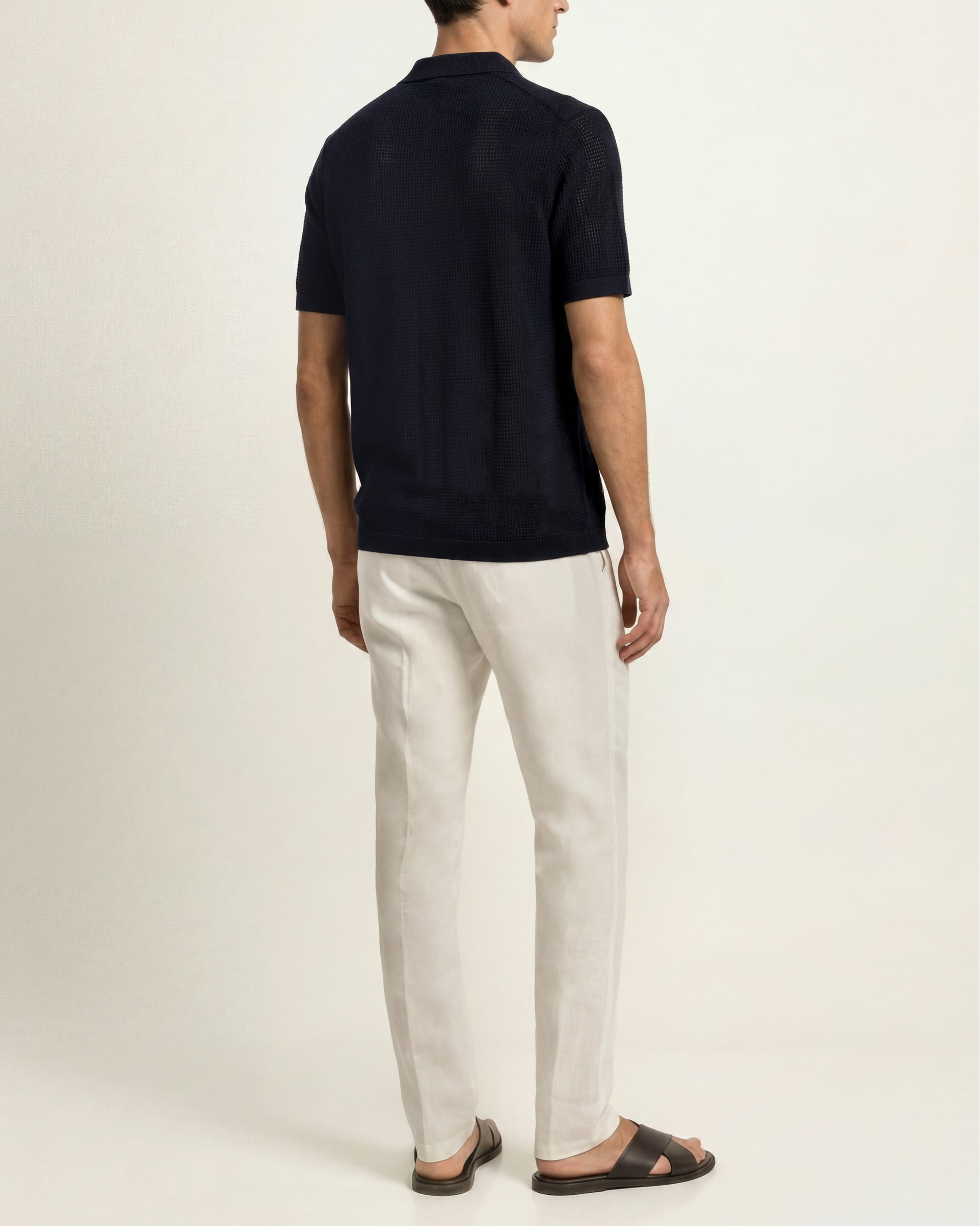 A man shown from the back wearing a dark navy short-sleeve polo shirt, off-white trousers, and dark brown sandals, standing against a plain light beige studio background.