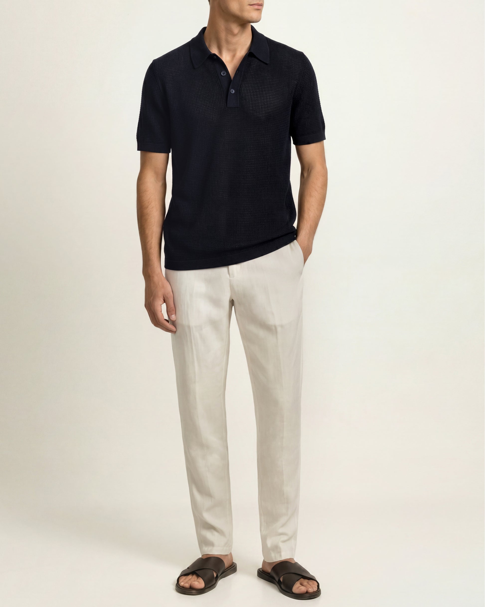 A man wearing a dark navy short-sleeve polo shirt, off-white linen trousers, and dark brown slide sandals, standing against a plain light beige studio background.