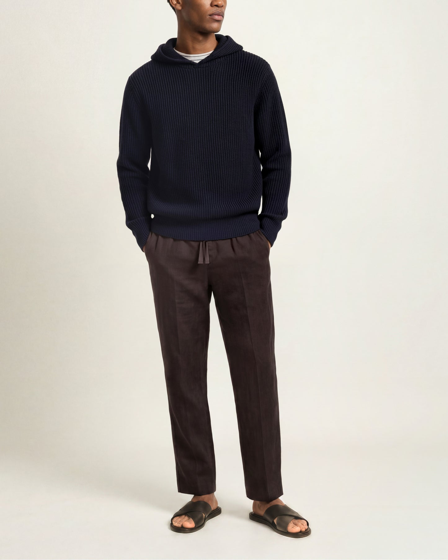 A man wearing a dark navy ribbed knit hoodie, dark brown drawstring trousers, and dark brown slide sandals, standing against a plain light beige studio background.