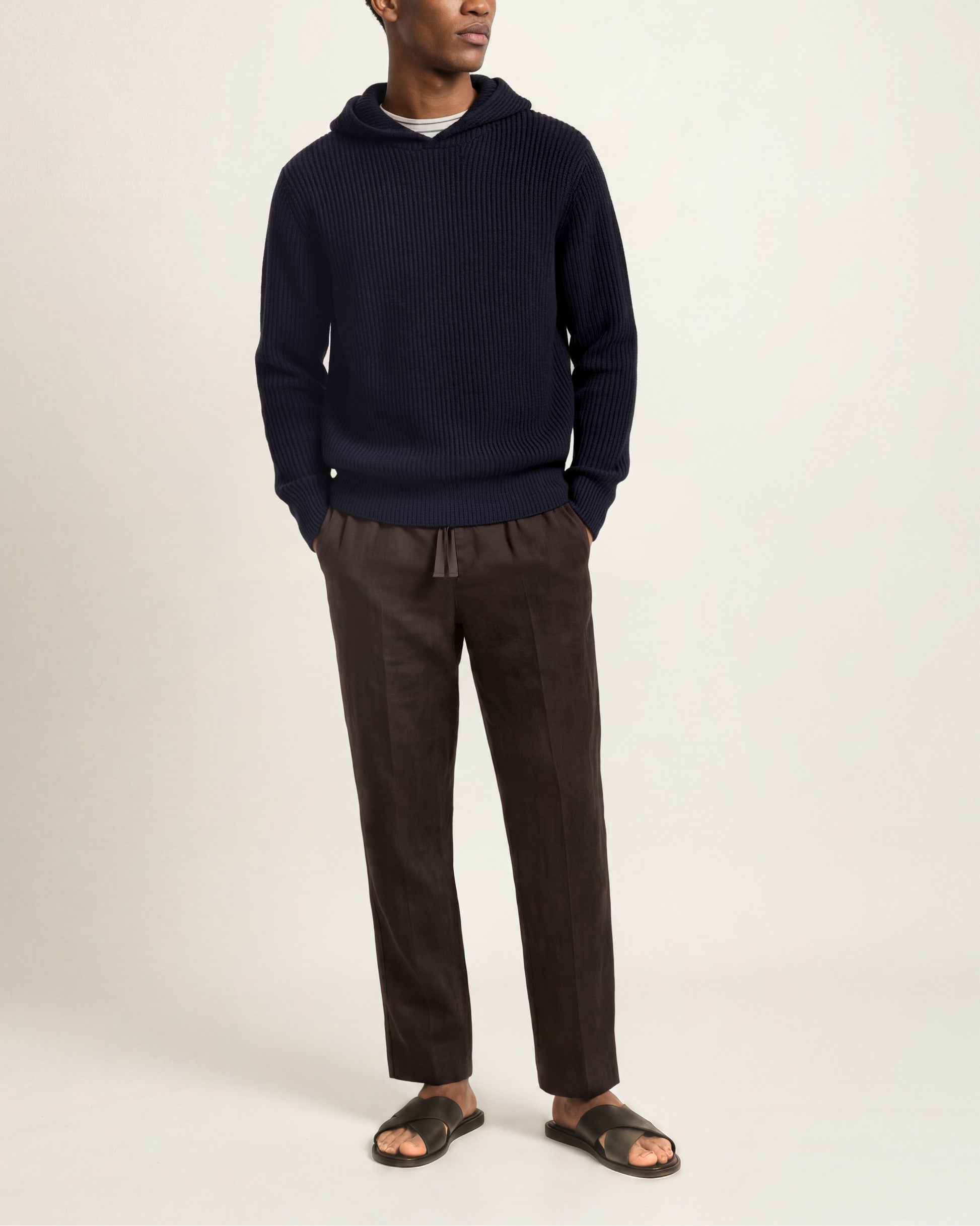 A man wearing a dark navy ribbed knit hoodie, dark brown drawstring trousers, and dark brown slide sandals, standing against a plain light beige studio background.
