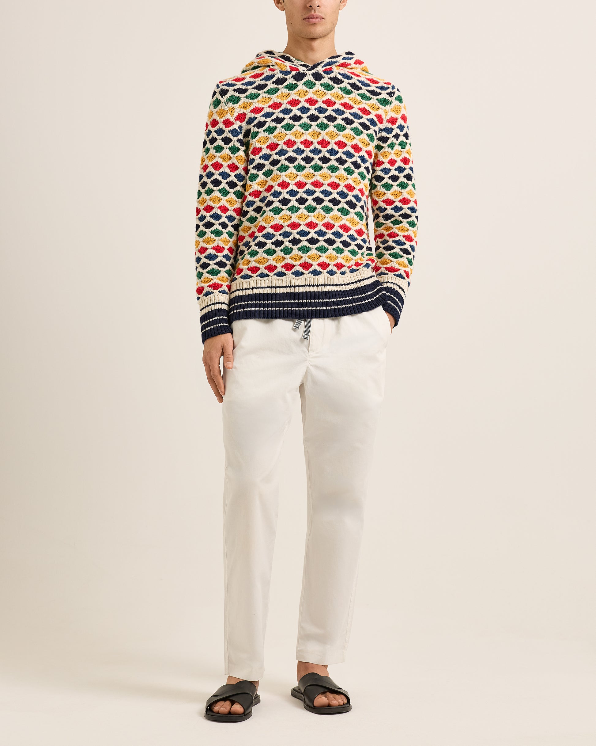 Person wearing a colorful patterned sweater and white pants on a beige background