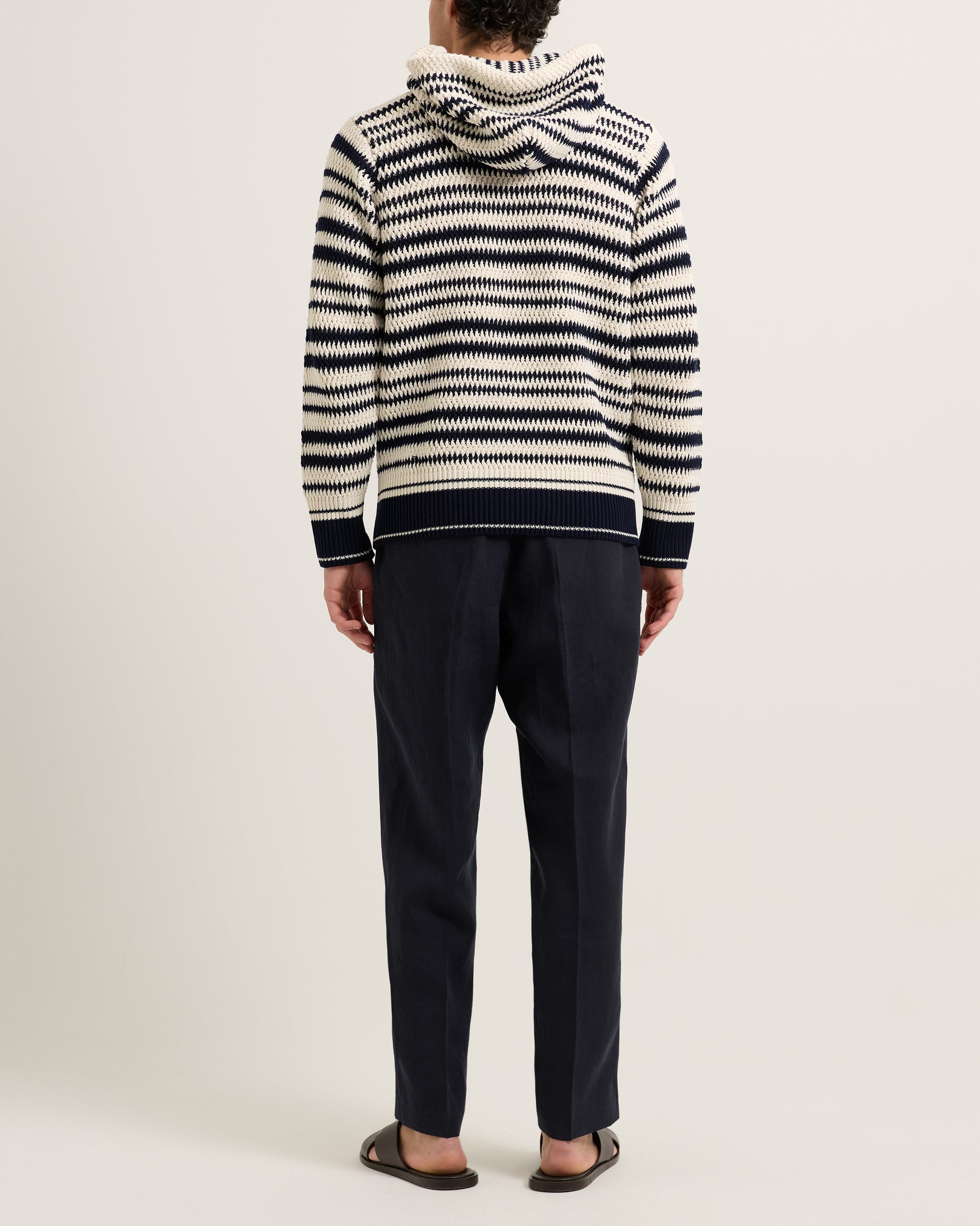 Man wearing knitted striped hoodie with navy trousers on a white background