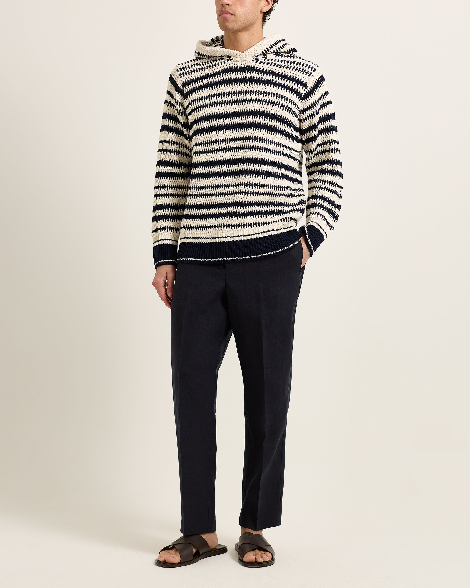 Man wearing knitted striped hoodie with navy trousers on a white background