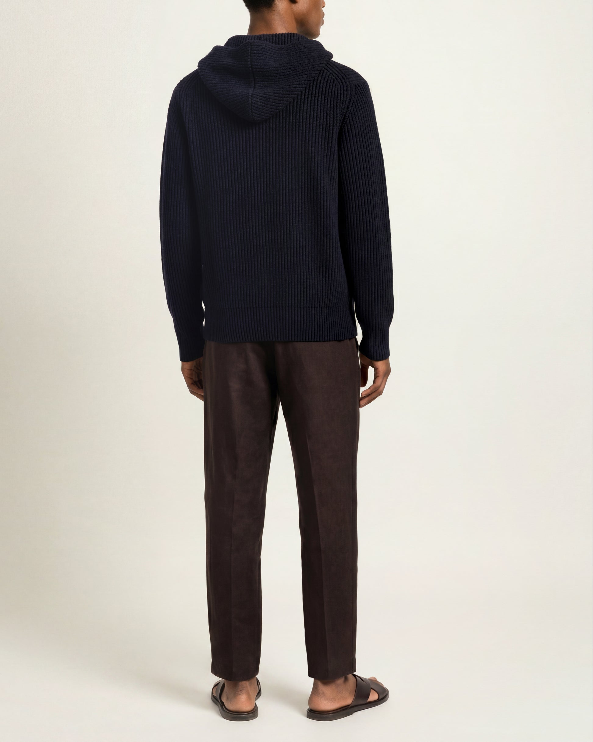 A man viewed from the back wearing a dark navy ribbed knit hooded sweater, dark brown trousers, and dark sandals, standing against a plain off-white studio background.