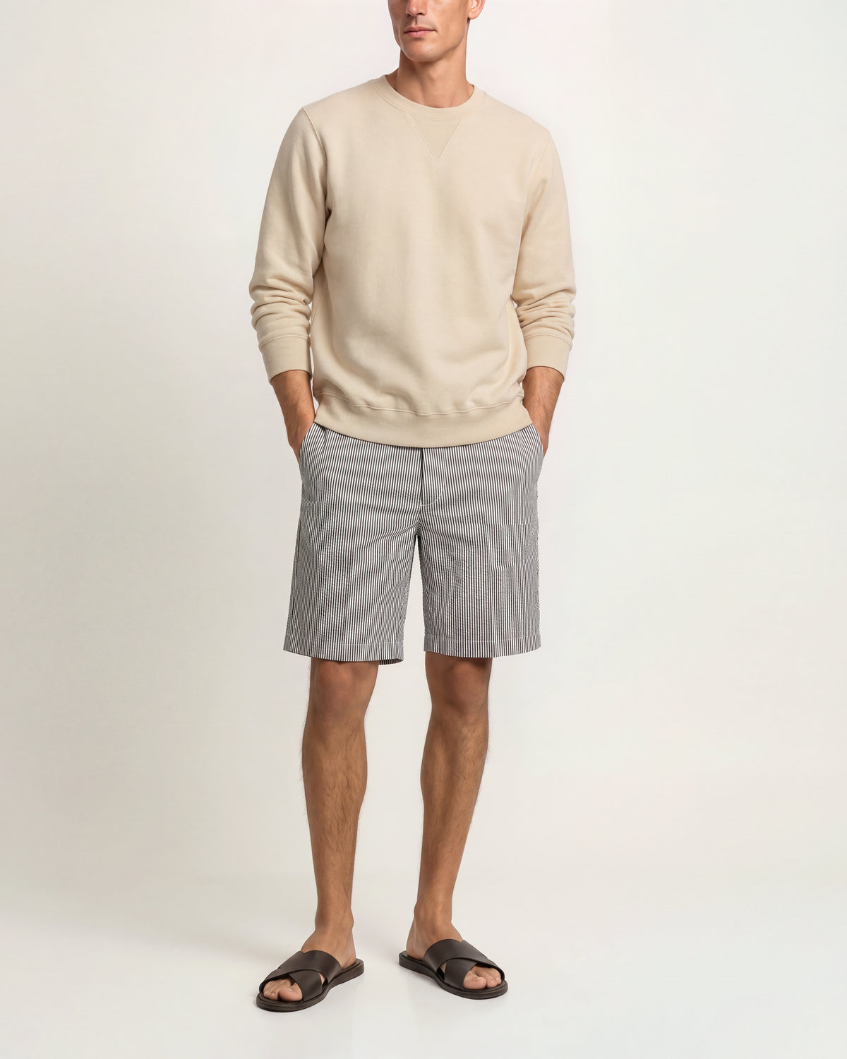 A man modeling a light beige crewneck sweatshirt and light gray shorts, paired with dark brown slide sandals, against a plain off-white studio background.