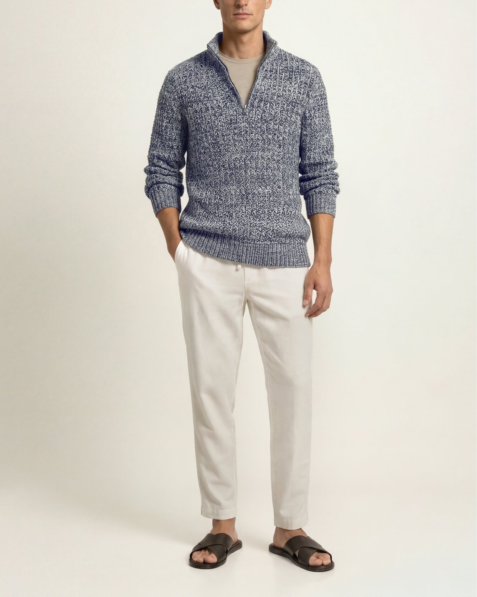 A man wearing a textured blue and white quarter-zip knit sweater, off-white trousers, and dark brown slide sandals, standing against a plain light beige studio background.