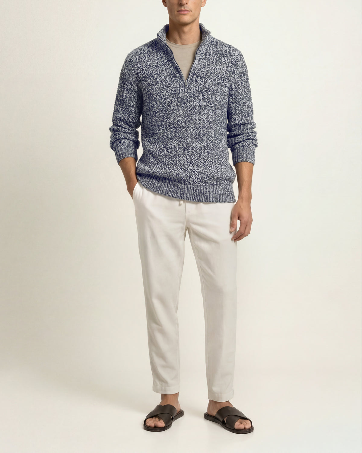 A man wearing a textured blue and white quarter-zip knit sweater, off-white trousers, and dark brown slide sandals, standing against a plain light beige studio background.