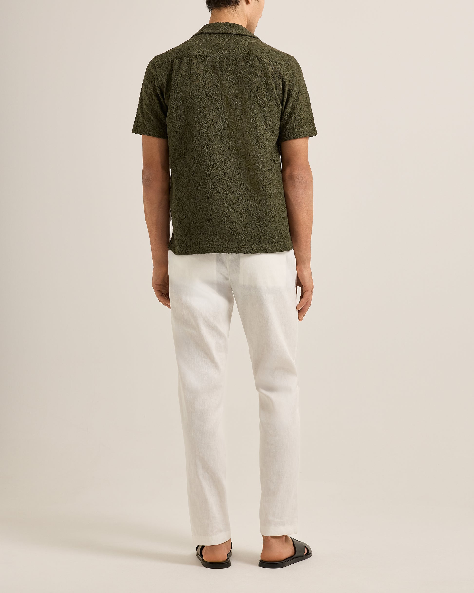 Person wearing a green patterned shirt and white pants on a beige background