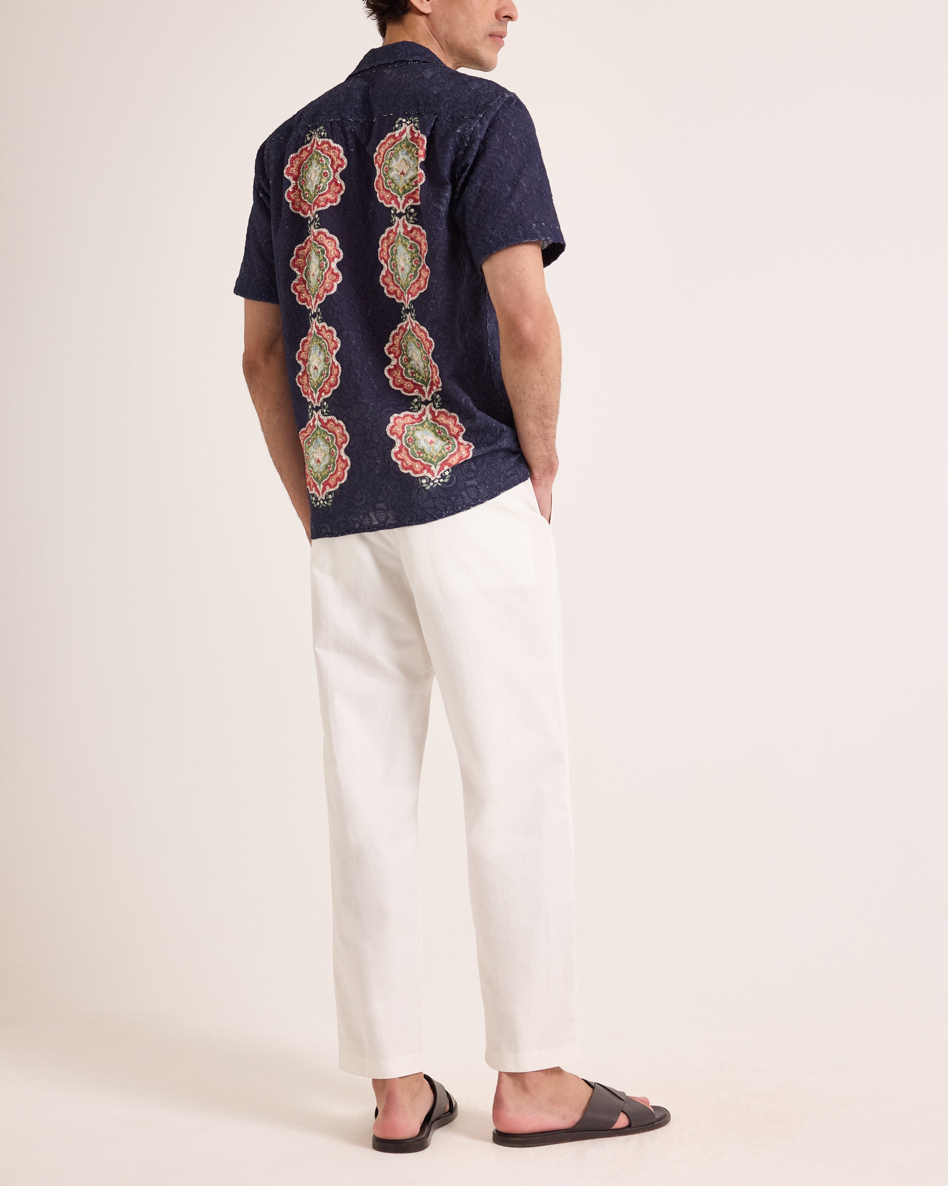 Man wearing patterned broderie shirt with white trousers on a plain background
