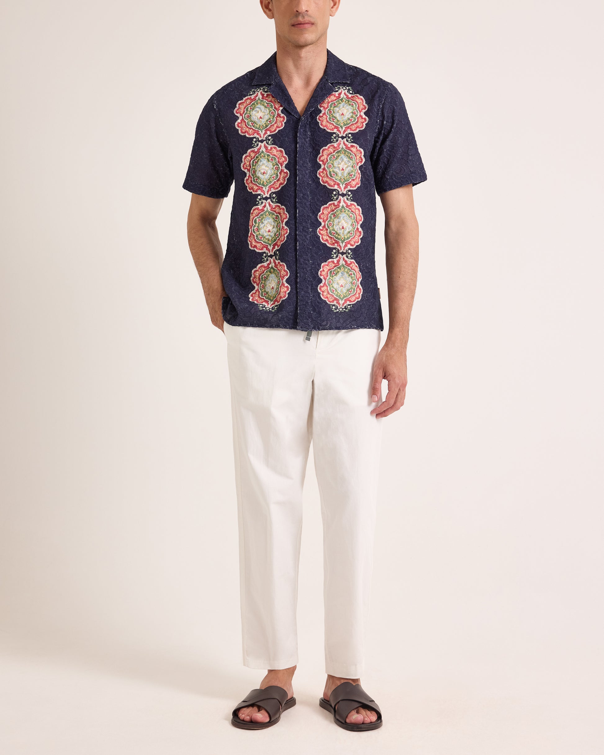 Man wearing patterned broderie shirt with white trousers on a plain background