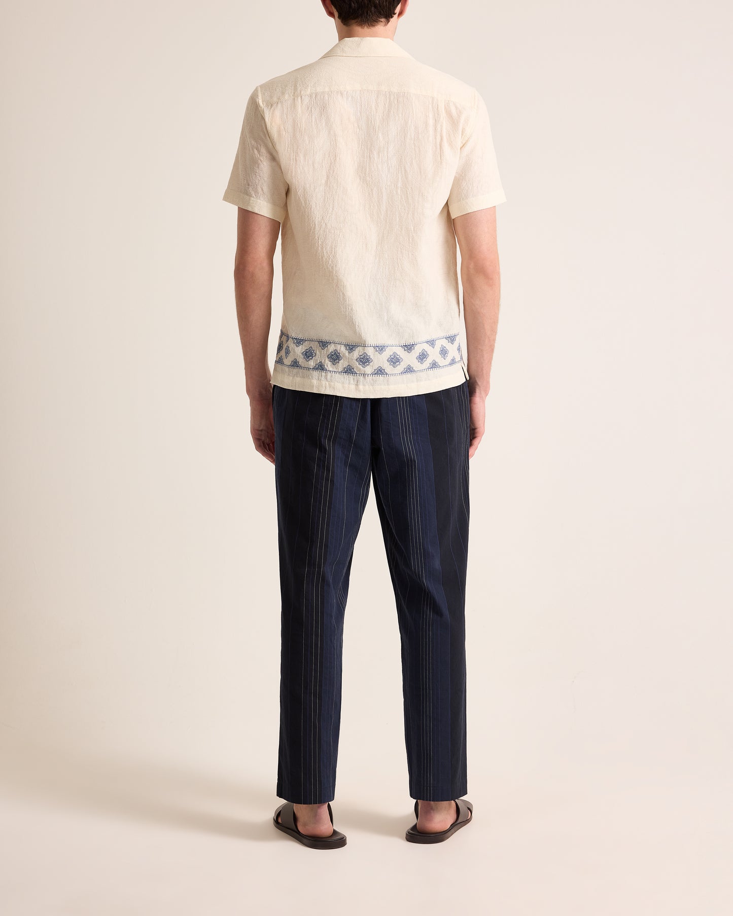 Person wearing a beige shirt with a geometric pattern and dark blue pants on a plain background