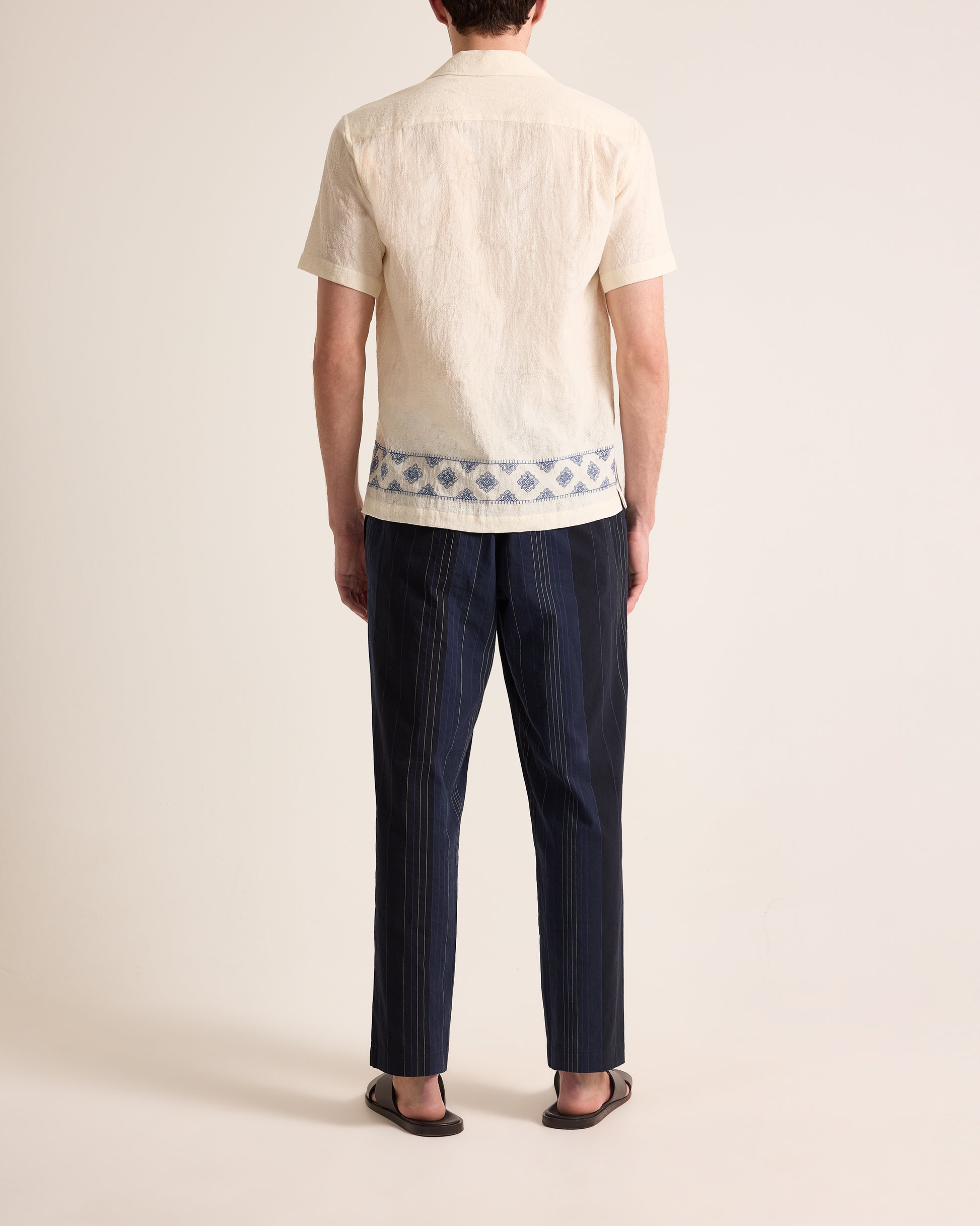 Person wearing a beige shirt with a geometric pattern and dark blue pants on a plain background