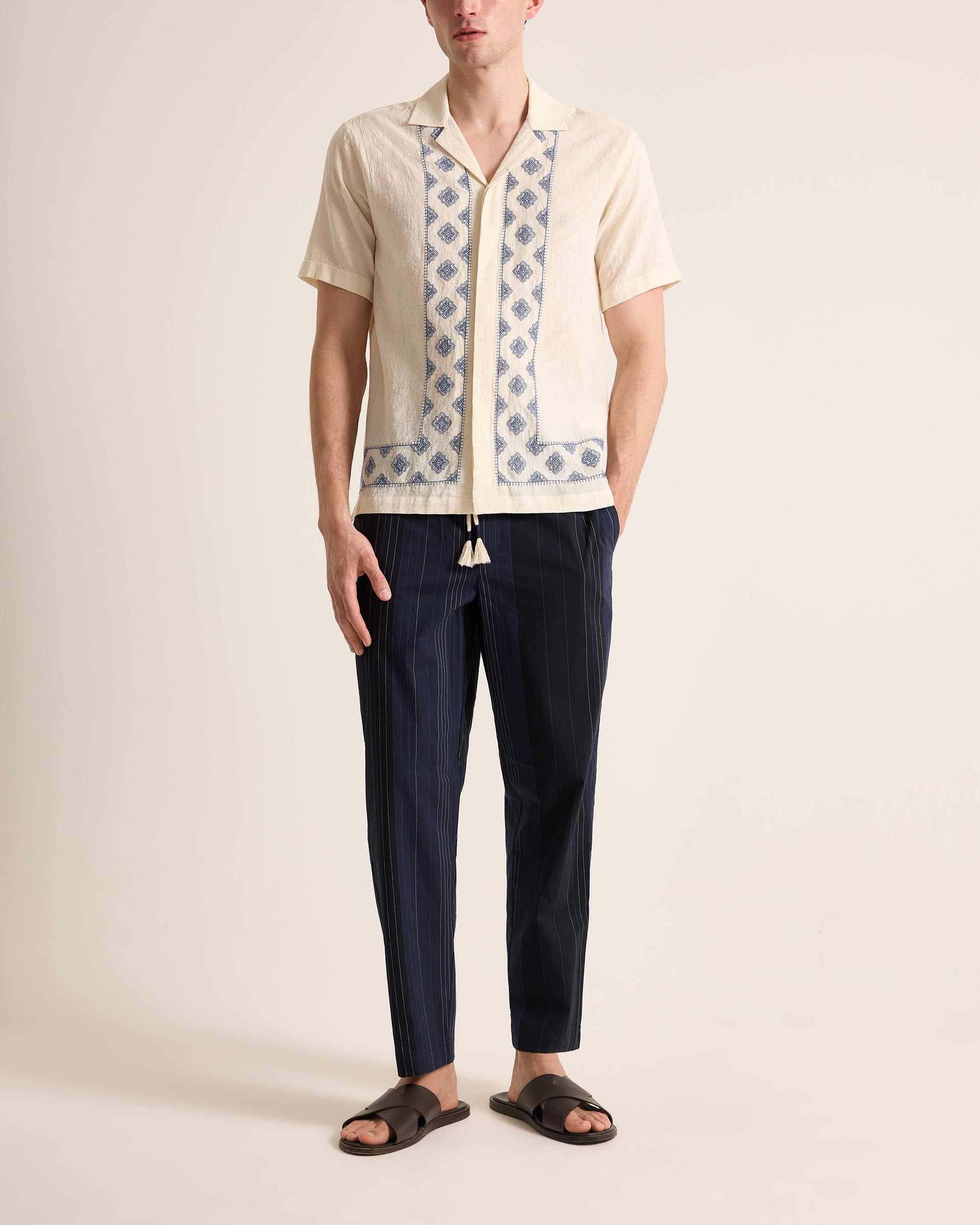 Person wearing a patterned shirt and dark pants on a plain background