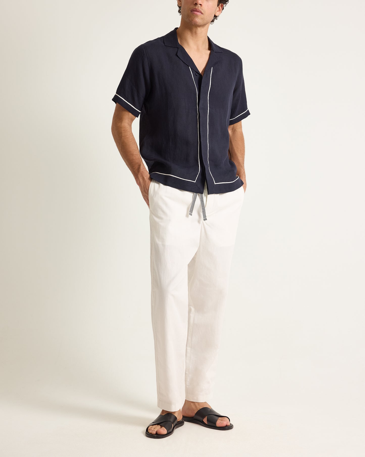 Man wearing a navy shirt with white trim and white pants on a plain background