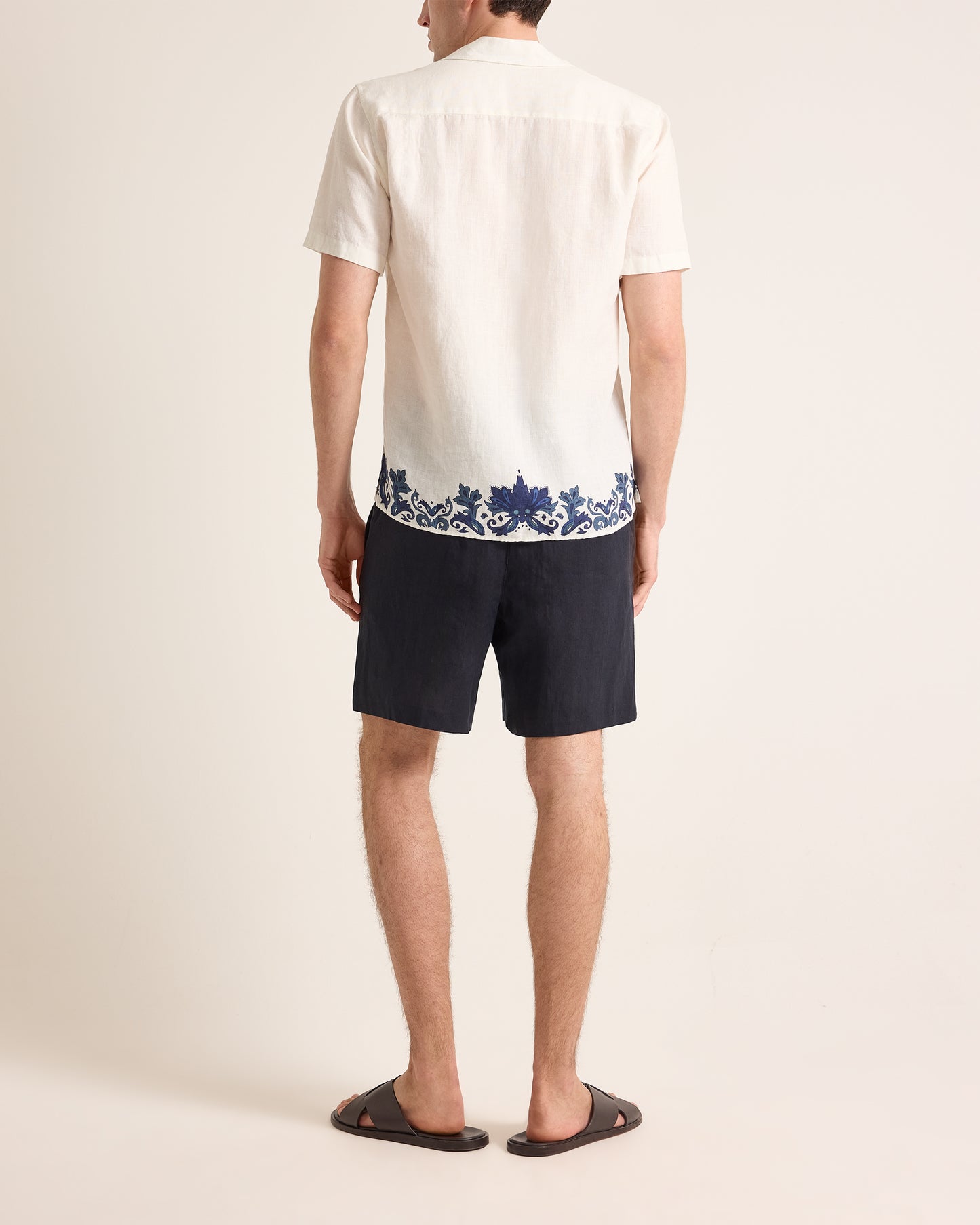 Person wearing a white shirt with blue floral embroidery and dark shorts on a plain background