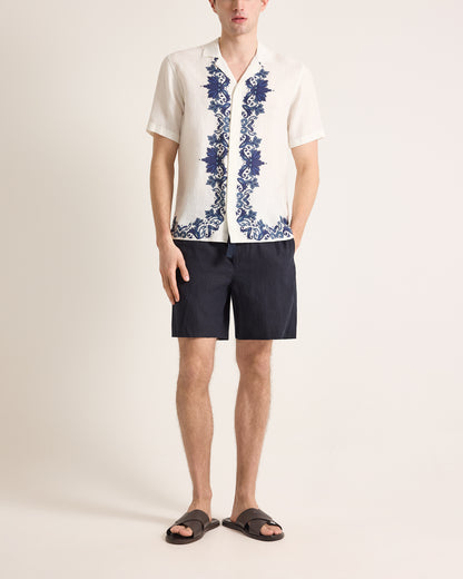 Person wearing a white shirt with blue floral embroidery and black shorts on a plain background