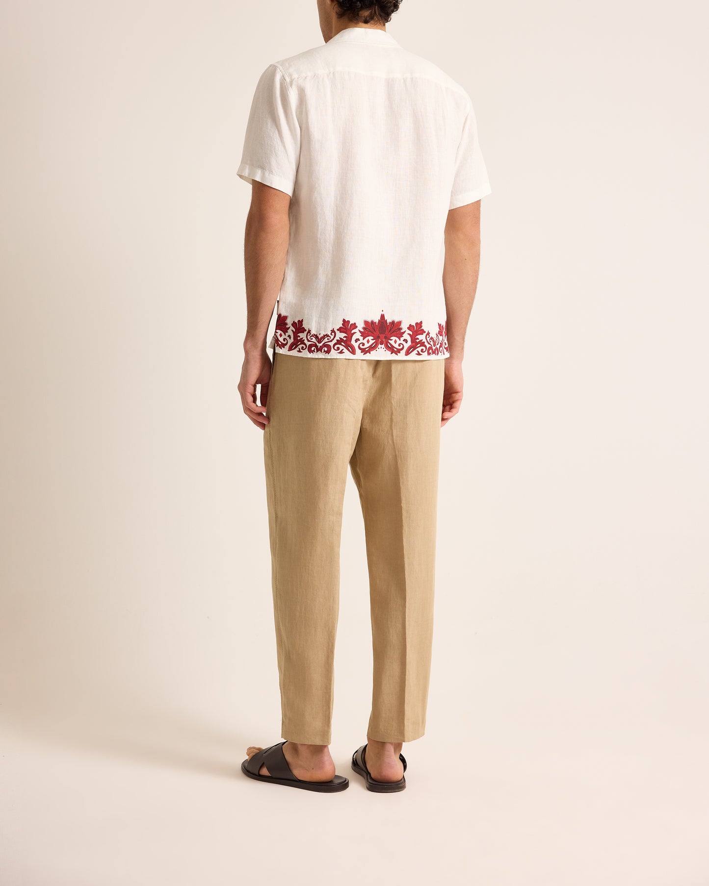 Person wearing a white shirt with red floral embroidery and beige pants on a plain background