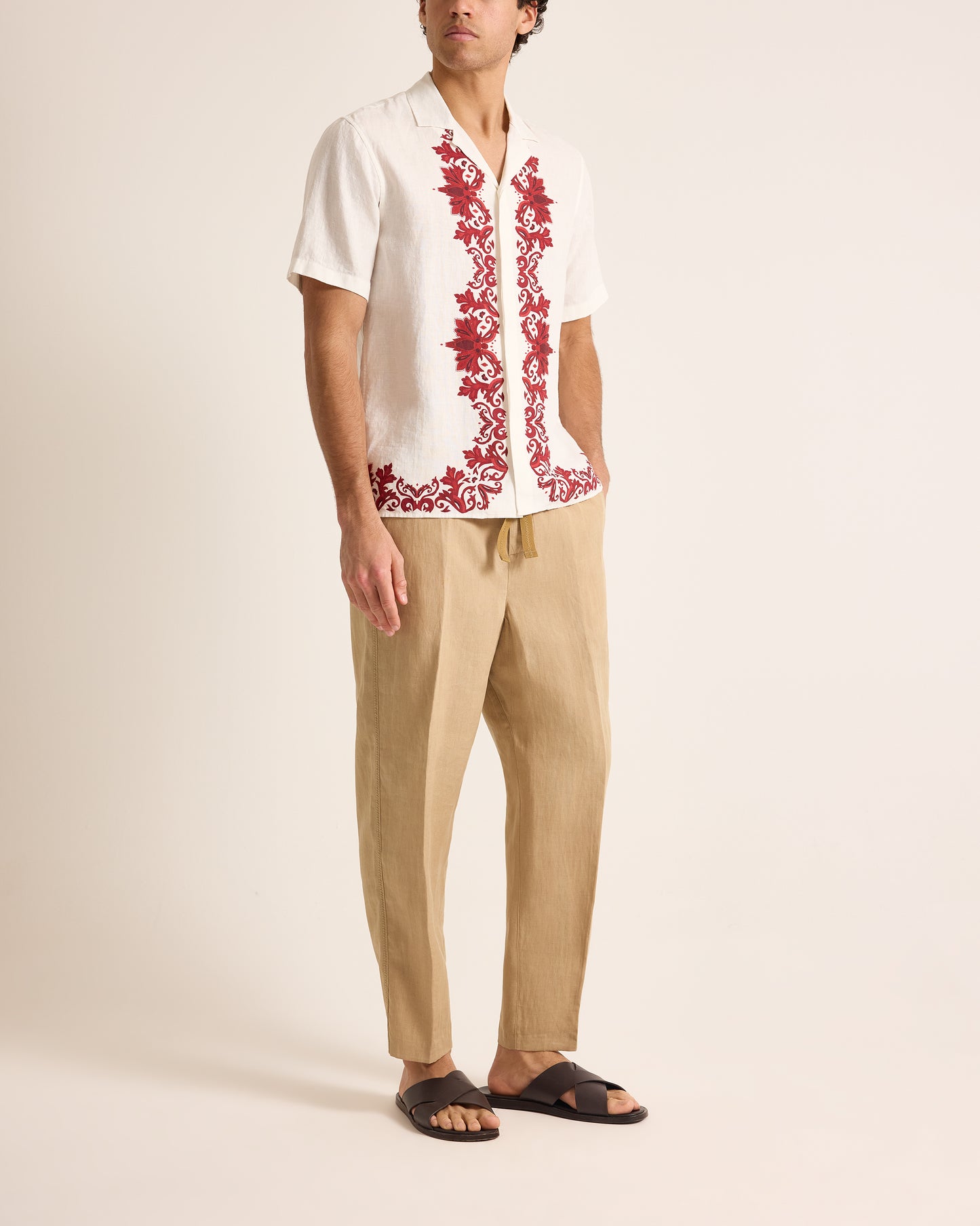 Man wearing a white shirt with red floral embroidery and beige pants on a plain background
