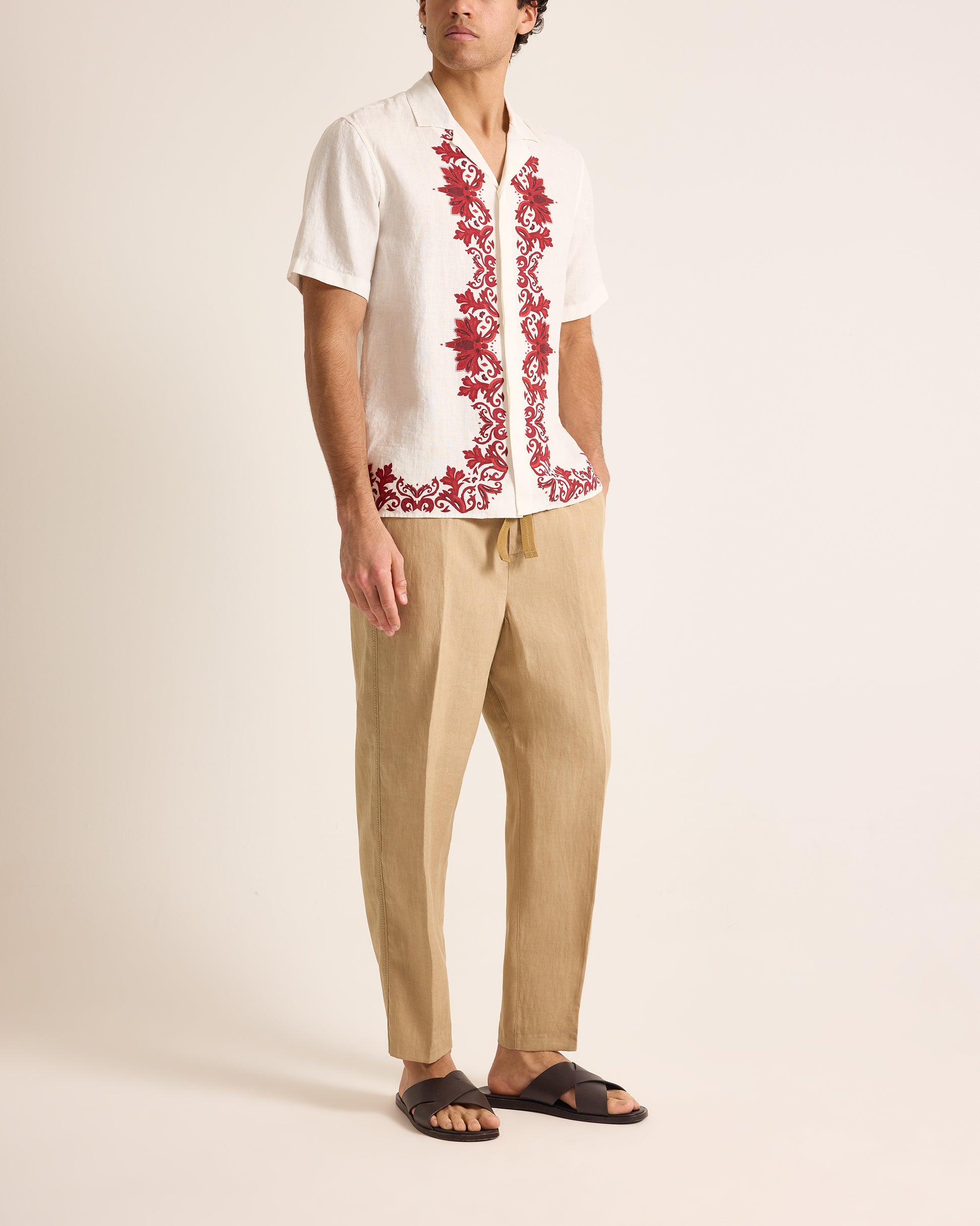 Man wearing a white shirt with red floral embroidery and beige pants on a plain background