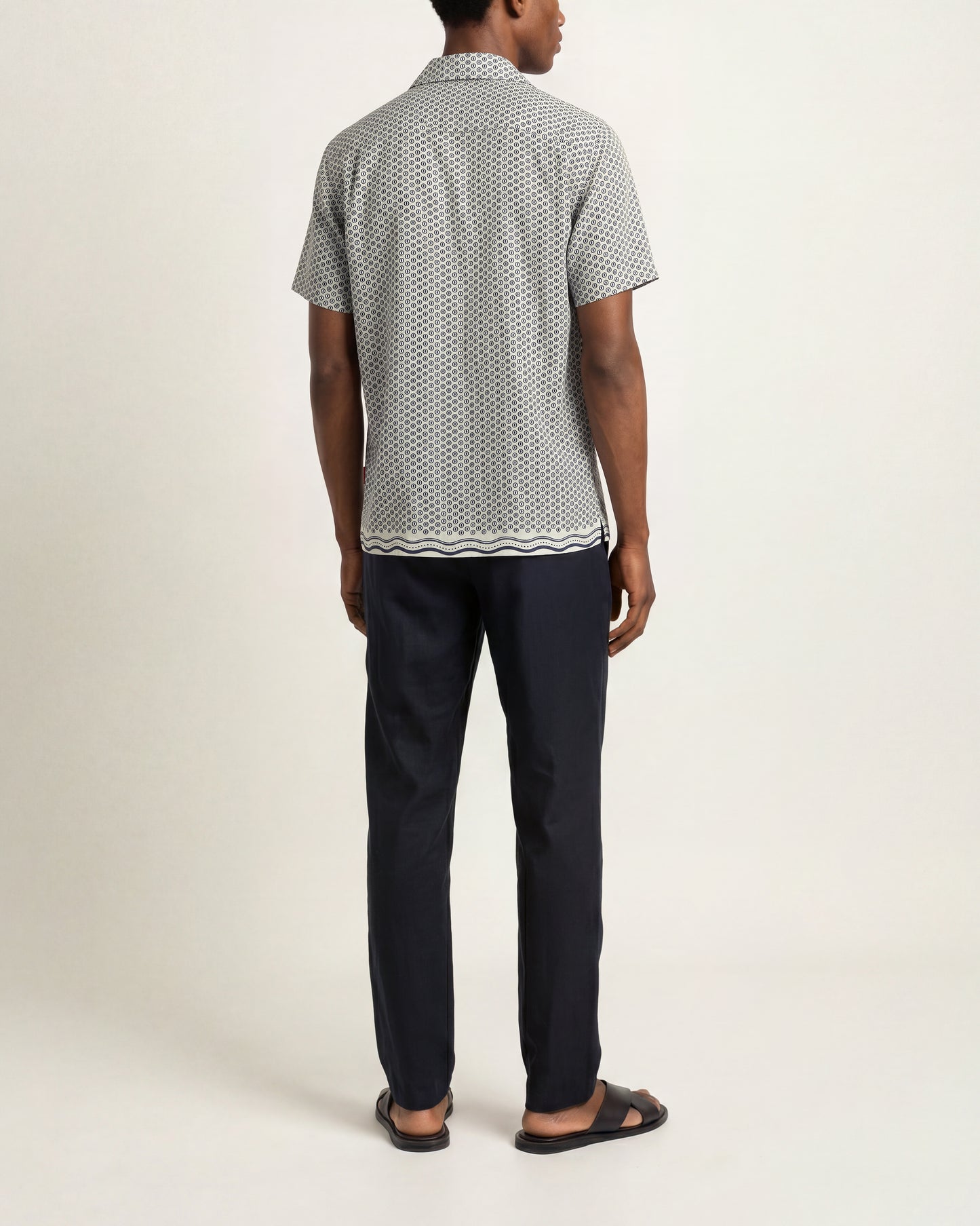 A man viewed from the back wearing a short-sleeve button-up shirt with a small white geometric pattern on a light gray base, paired with dark navy pants and black sandals, against a plain off-white studio background.