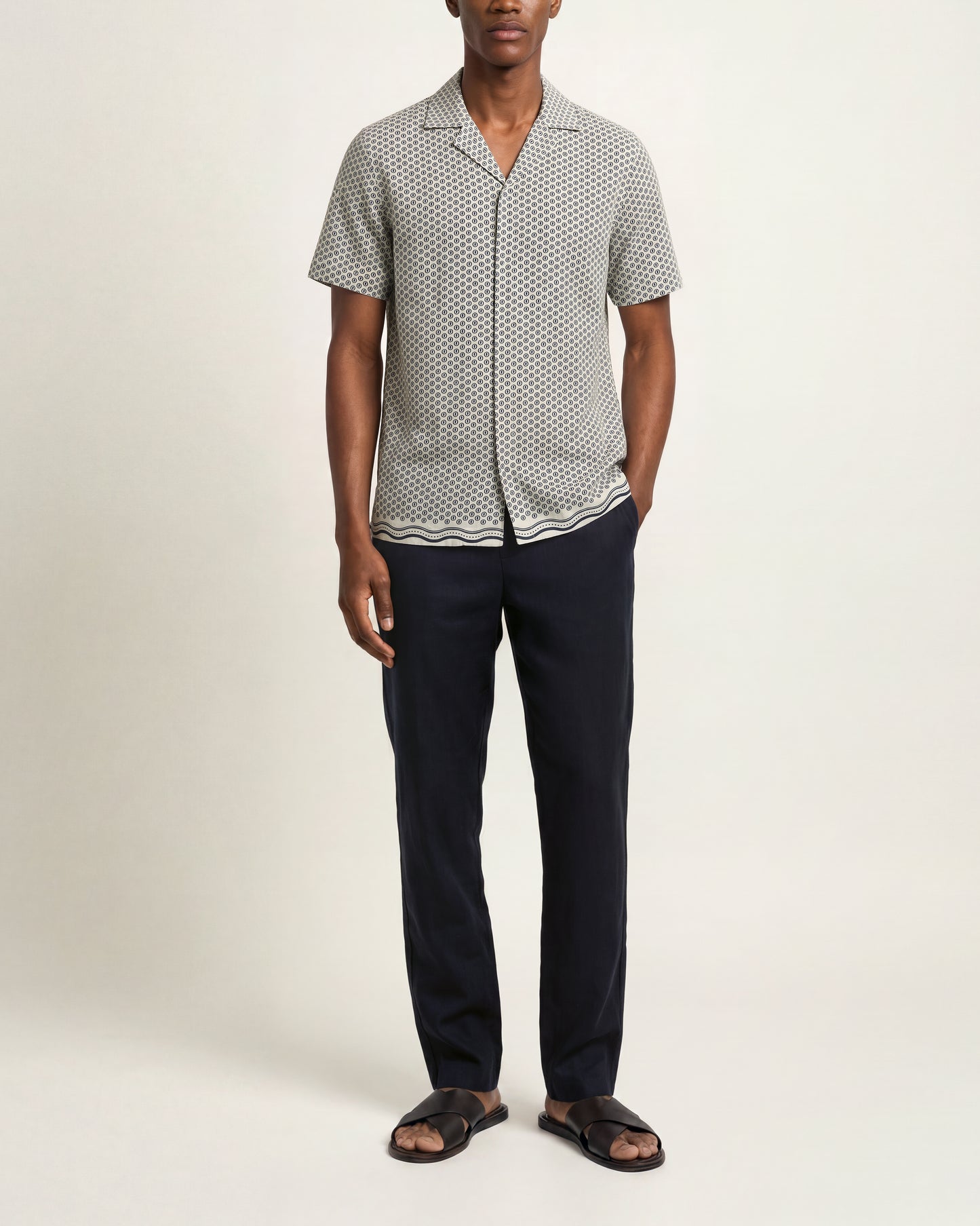 A man modeling a short-sleeve button-up shirt with a small white geometric pattern on a light gray background, paired with dark navy trousers and black slide sandals, against a plain off-white studio background.
