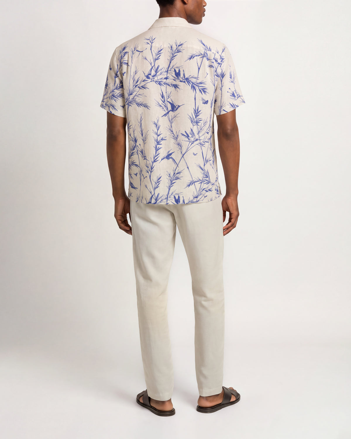 A man viewed from the back wearing a short-sleeve camp collar shirt with a blue tropical floral print on an off-white base, paired with off-white trousers and dark brown sandals, against a plain white studio background.