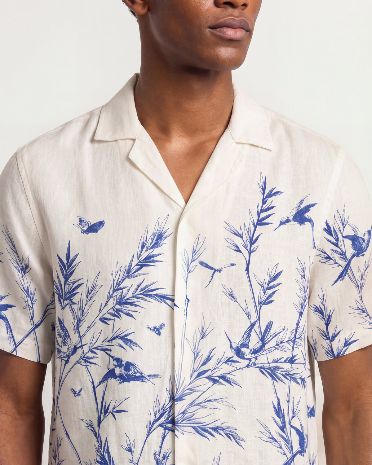 A close-up of a man wearing a short-sleeve cream-colored linen shirt with a blue bamboo and bird print, set against a plain light background.
