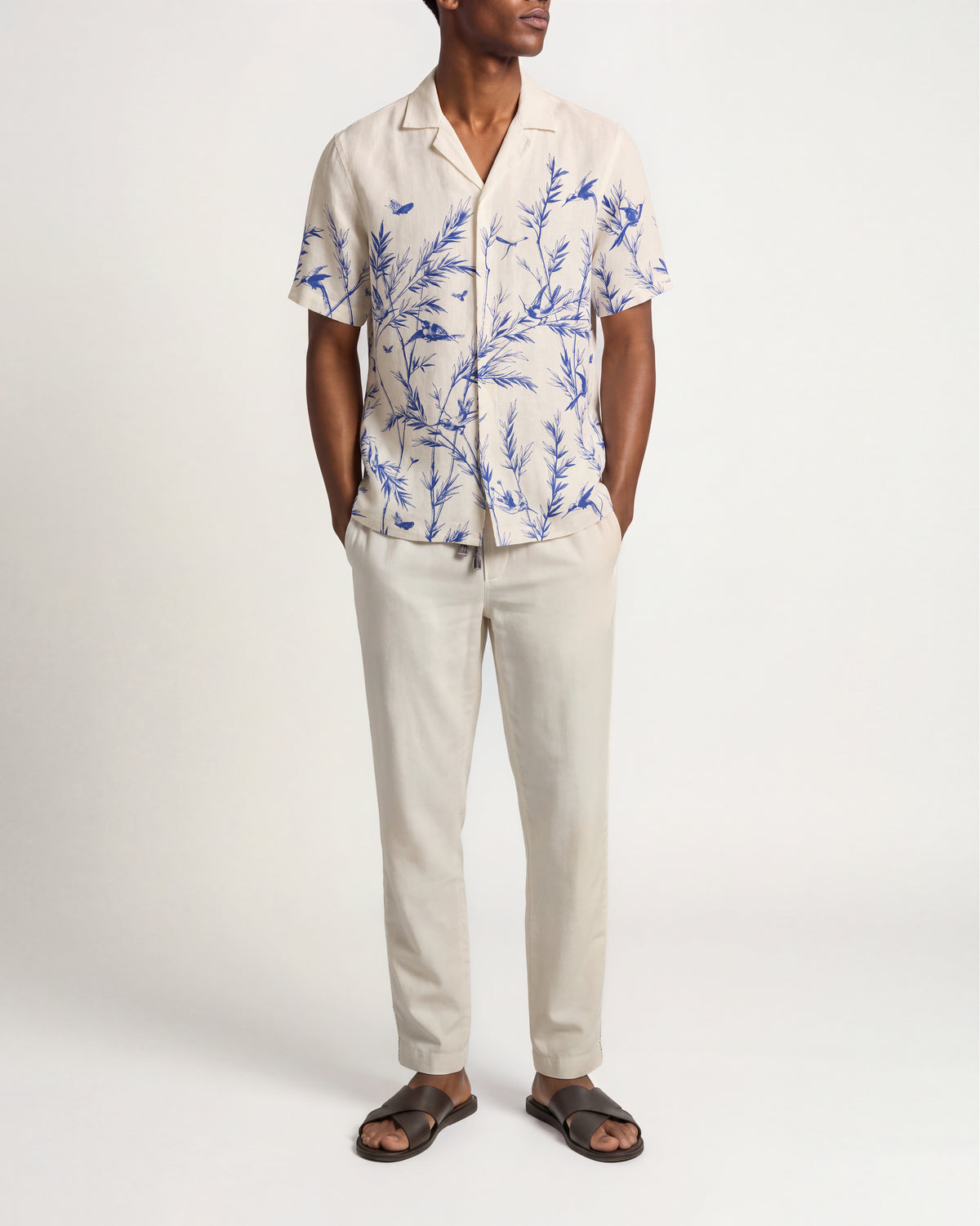 A man modeling a short-sleeve button-up shirt and matching trousers set. The shirt is cream-colored with a blue botanical and bird print, paired with solid cream trousers and dark brown slide sandals, shot against a plain white studio background.