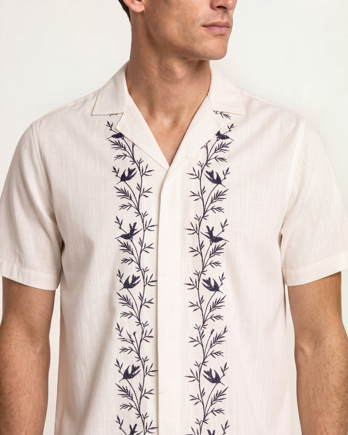 Close-up of a man wearing white short sleeved shirt with black embroidery detail on a white background